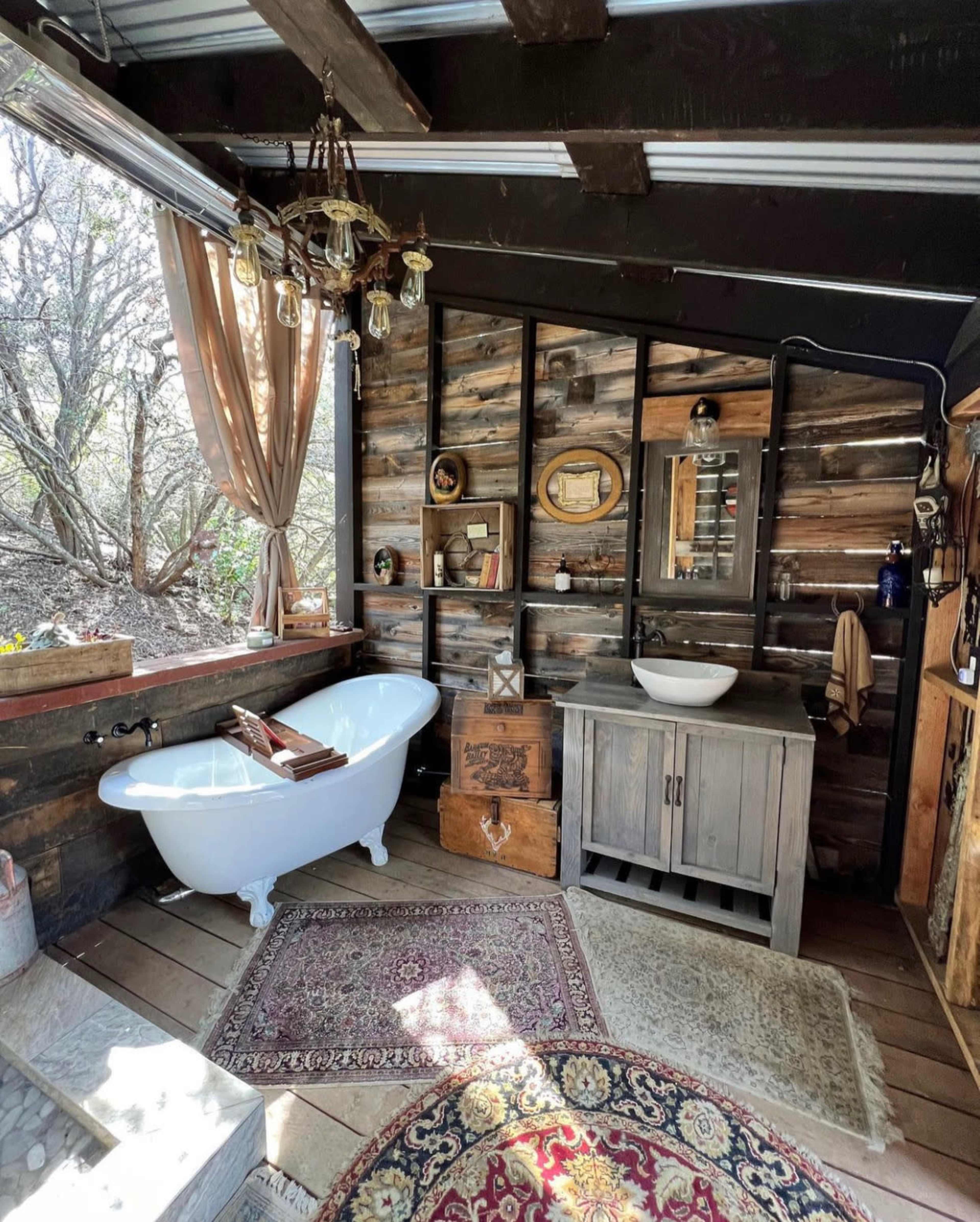 A rustic bathroom featuring a vintage clawfoot bathtub, a wooden sink cabinet, and various decorative items against a backdrop of wood paneling and natural light coming through a window.