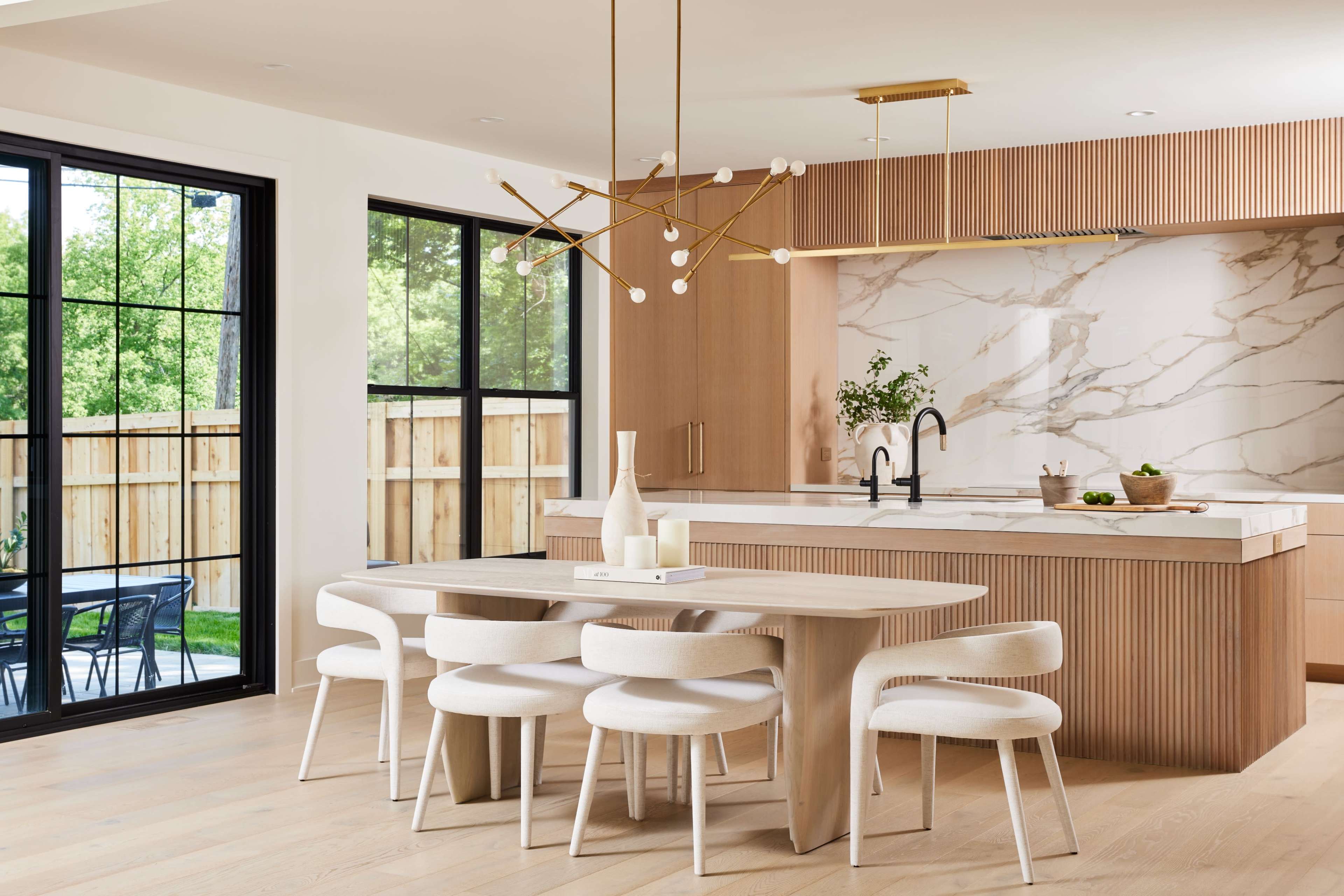 A modern kitchen features a wooden dining table surrounded by upholstered chairs, a minimalist chandelier, and a marble accent wall with a spacious island.