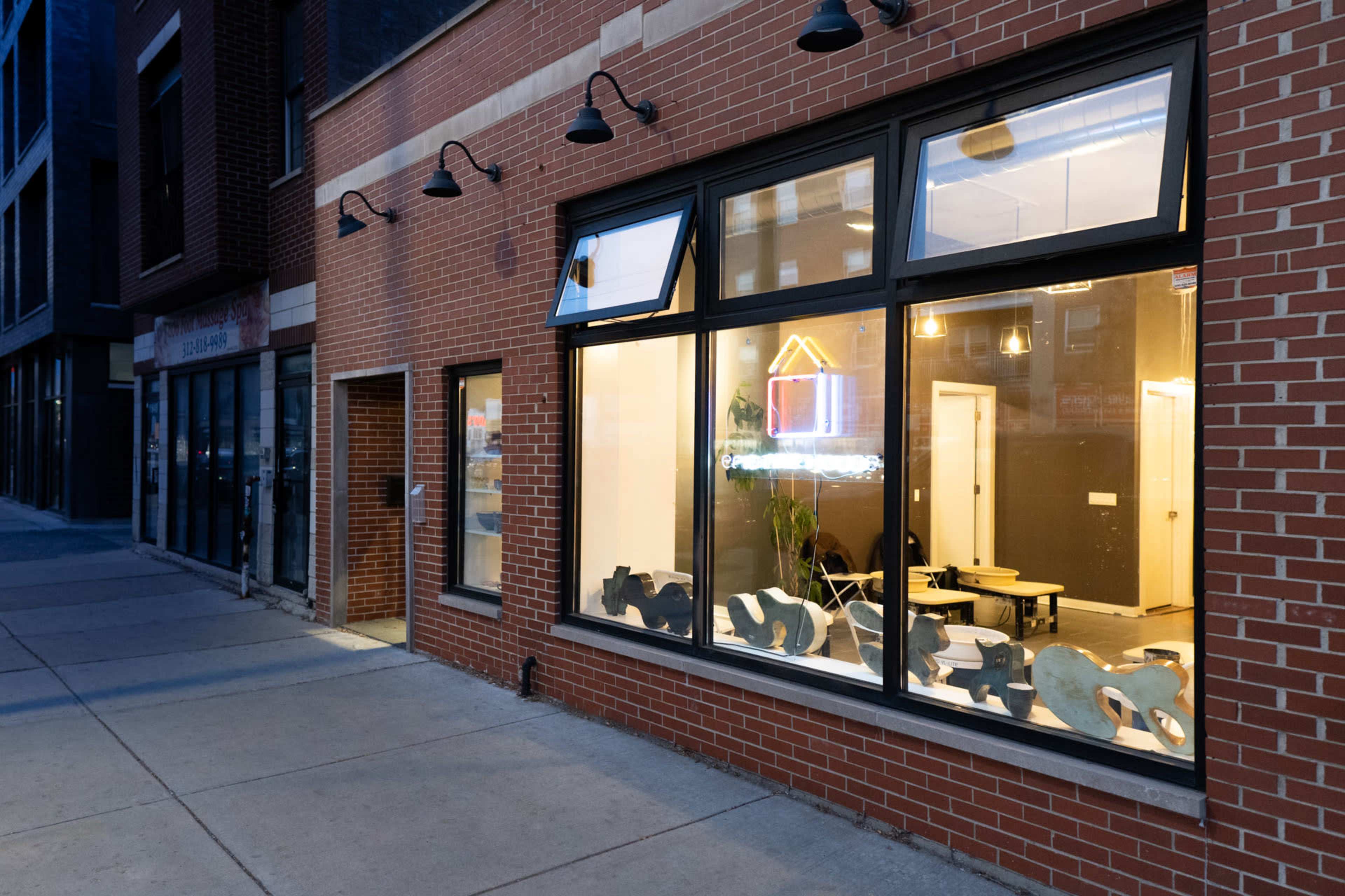 The exterior of a brick building with large windows illuminated at dusk, revealing a brightly lit interior with furniture and a neon sign.
