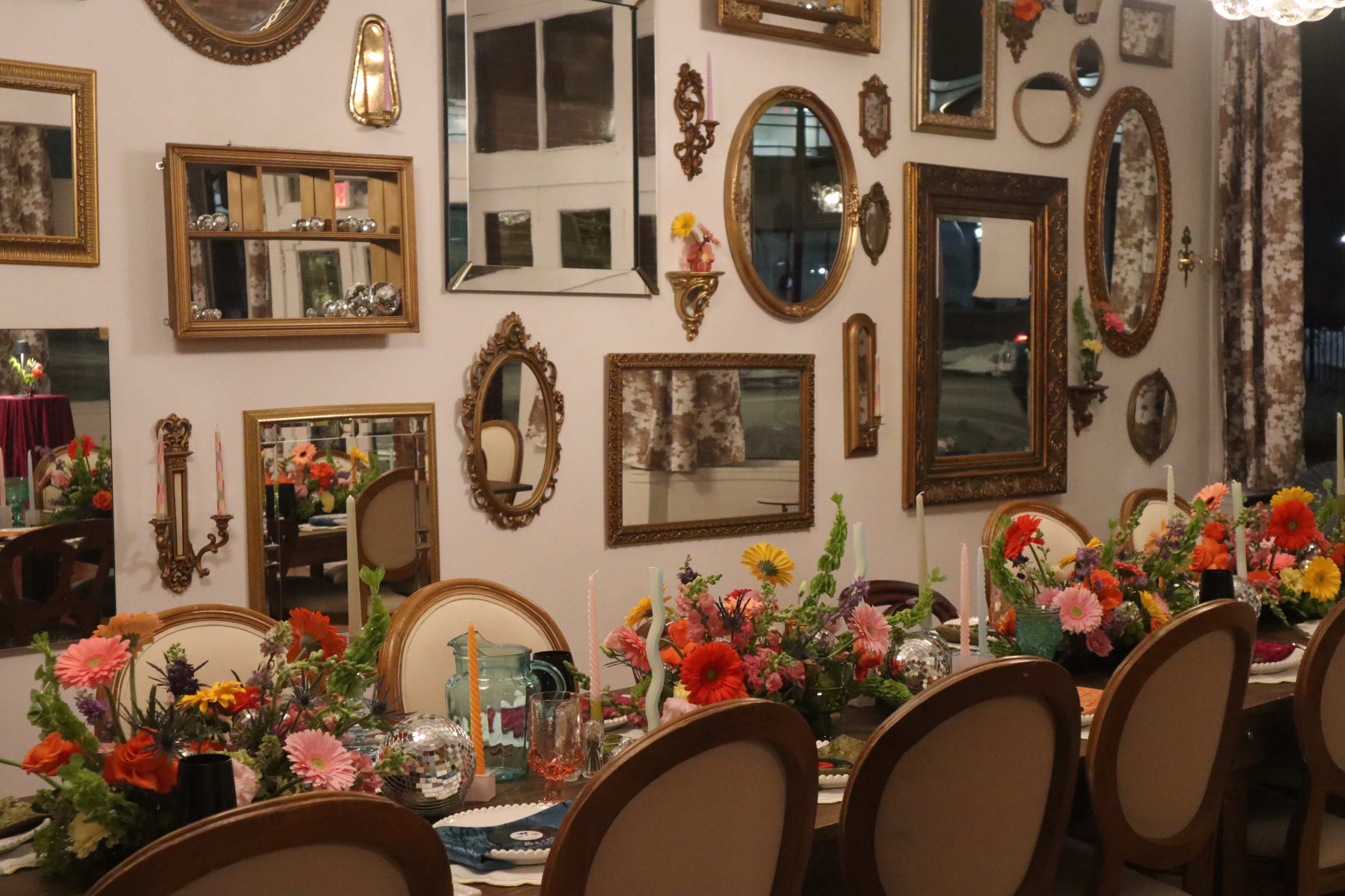 A long dining table adorned with vibrant flower arrangements sits against a wall lined with an assortment of decorative mirrors.