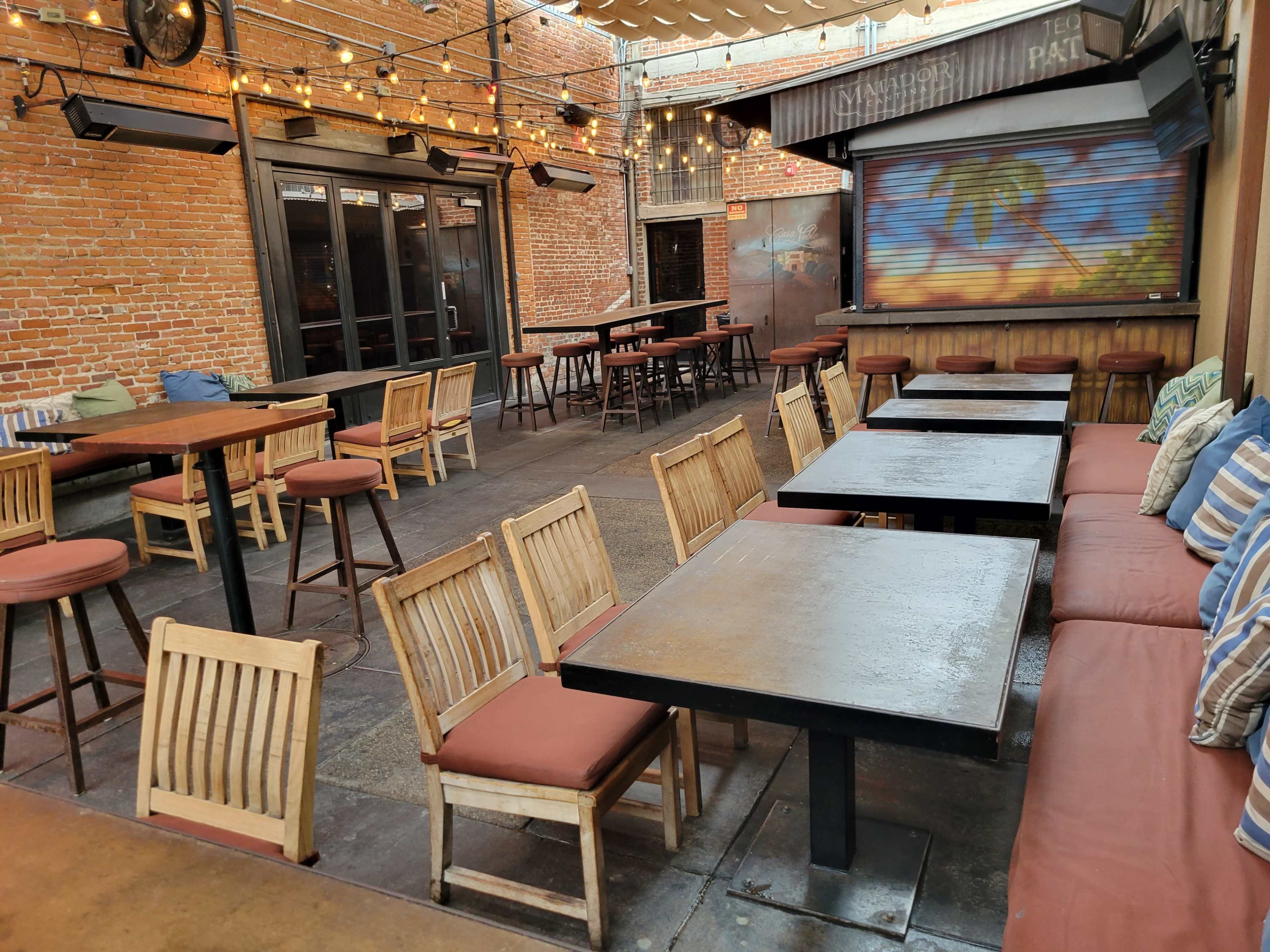 A patio area features wooden tables and chairs, with string lights overhead and a mural of a palm tree on the wall.
