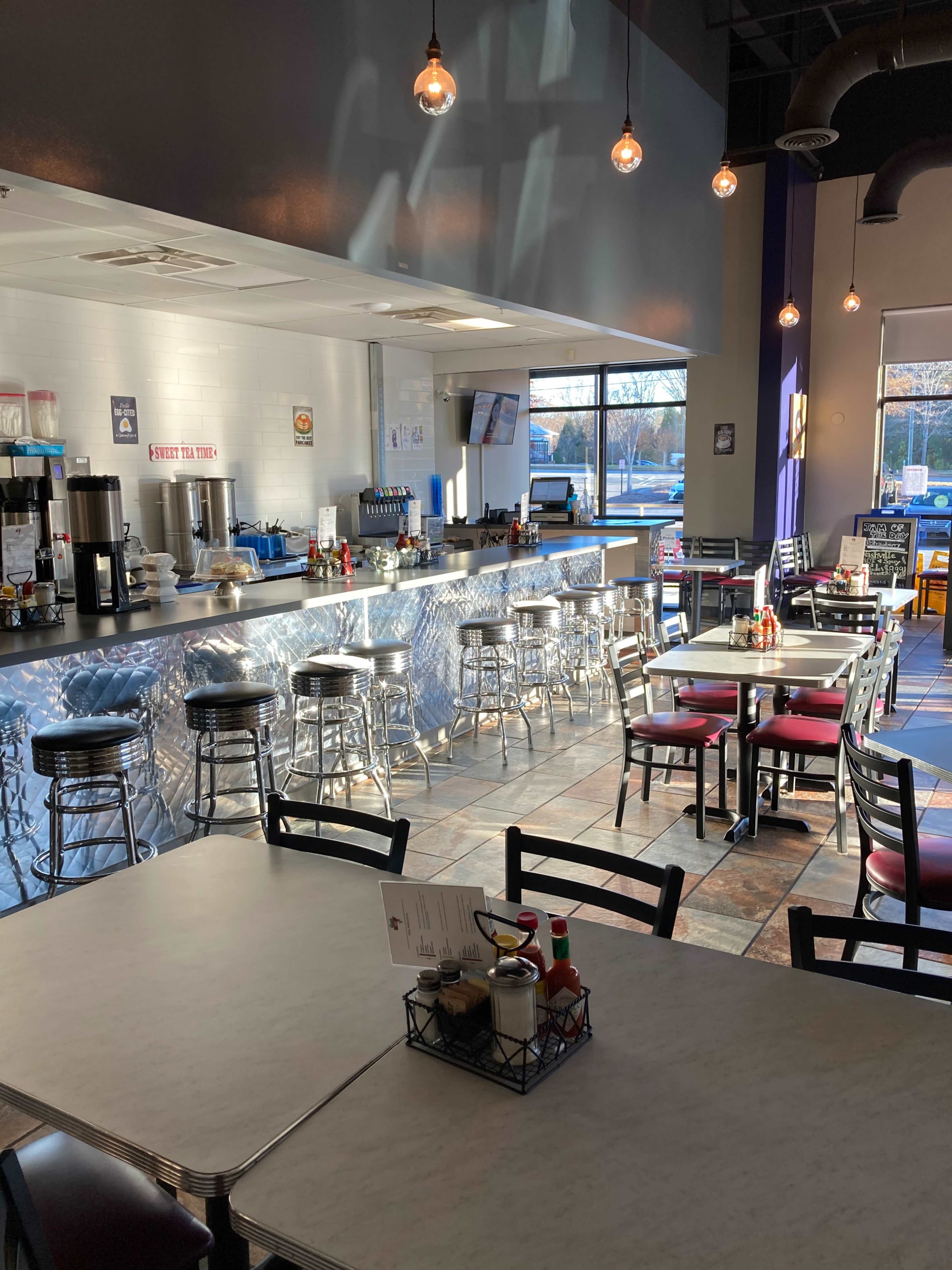 The image shows a modern diner interior with metal stools and tables, featuring a counter with drink stations and condiments visible on the tables.