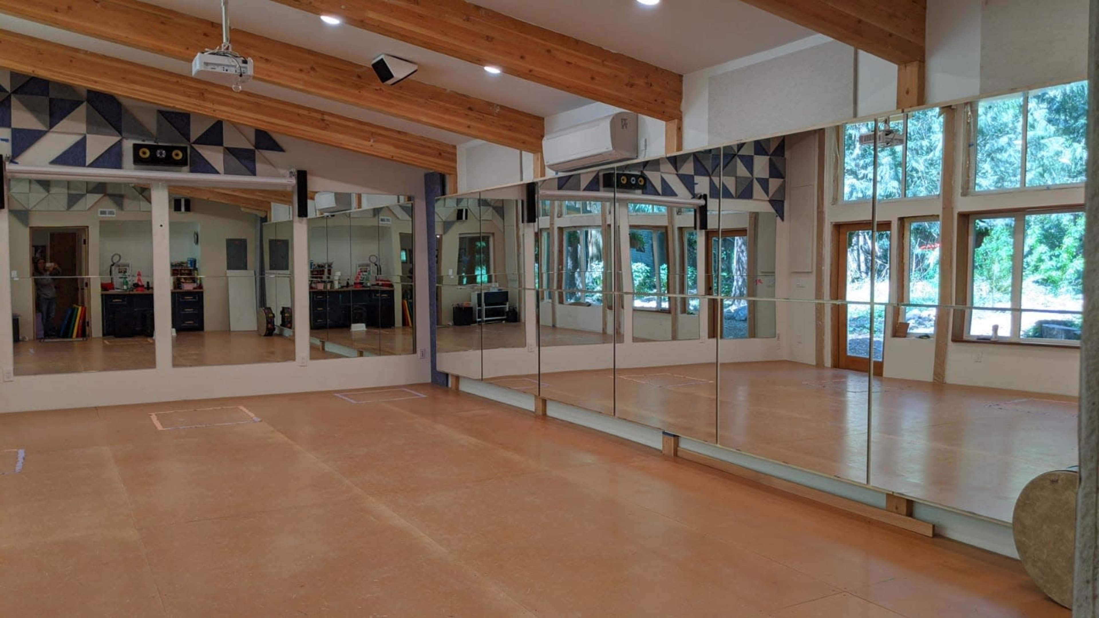 The image shows a spacious room with large mirrors on one wall, wooden beams on the ceiling, and windows letting in natural light.