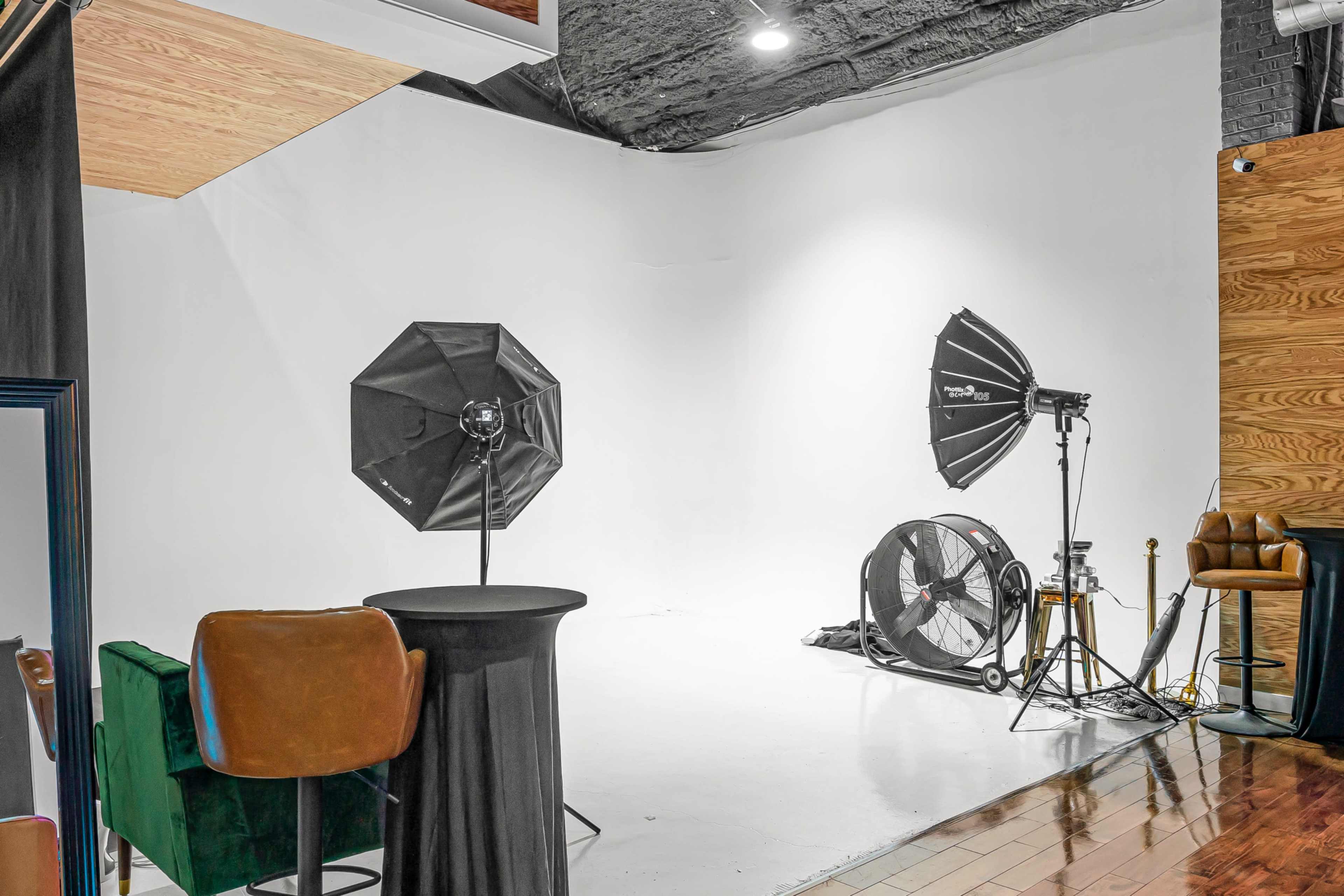 The image shows a photography studio with a white backdrop, two softbox lights, a fan, and various furniture pieces.