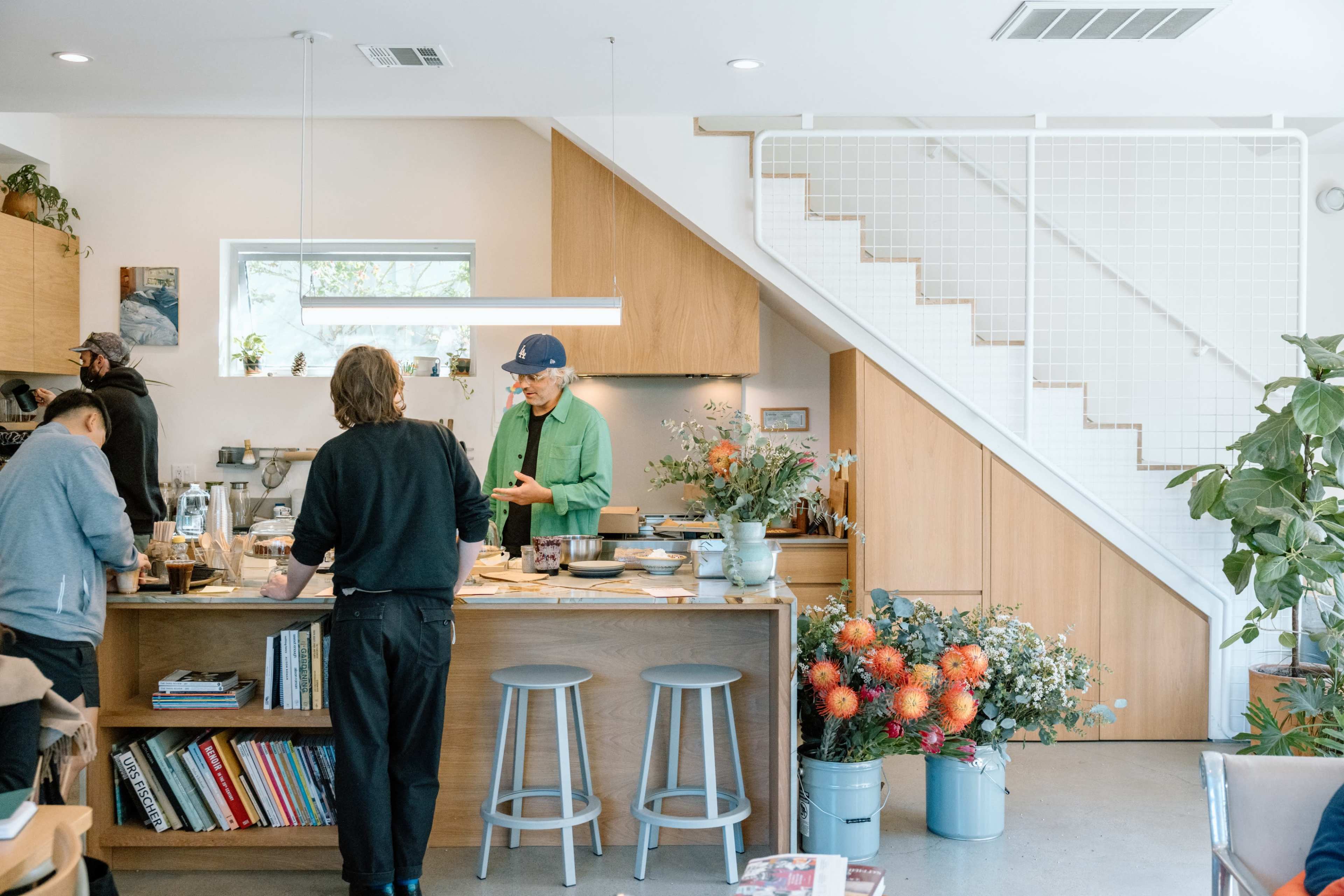 A group of people interact in a modern kitchen with a staircase in the background, surrounded by plants and kitchenware.