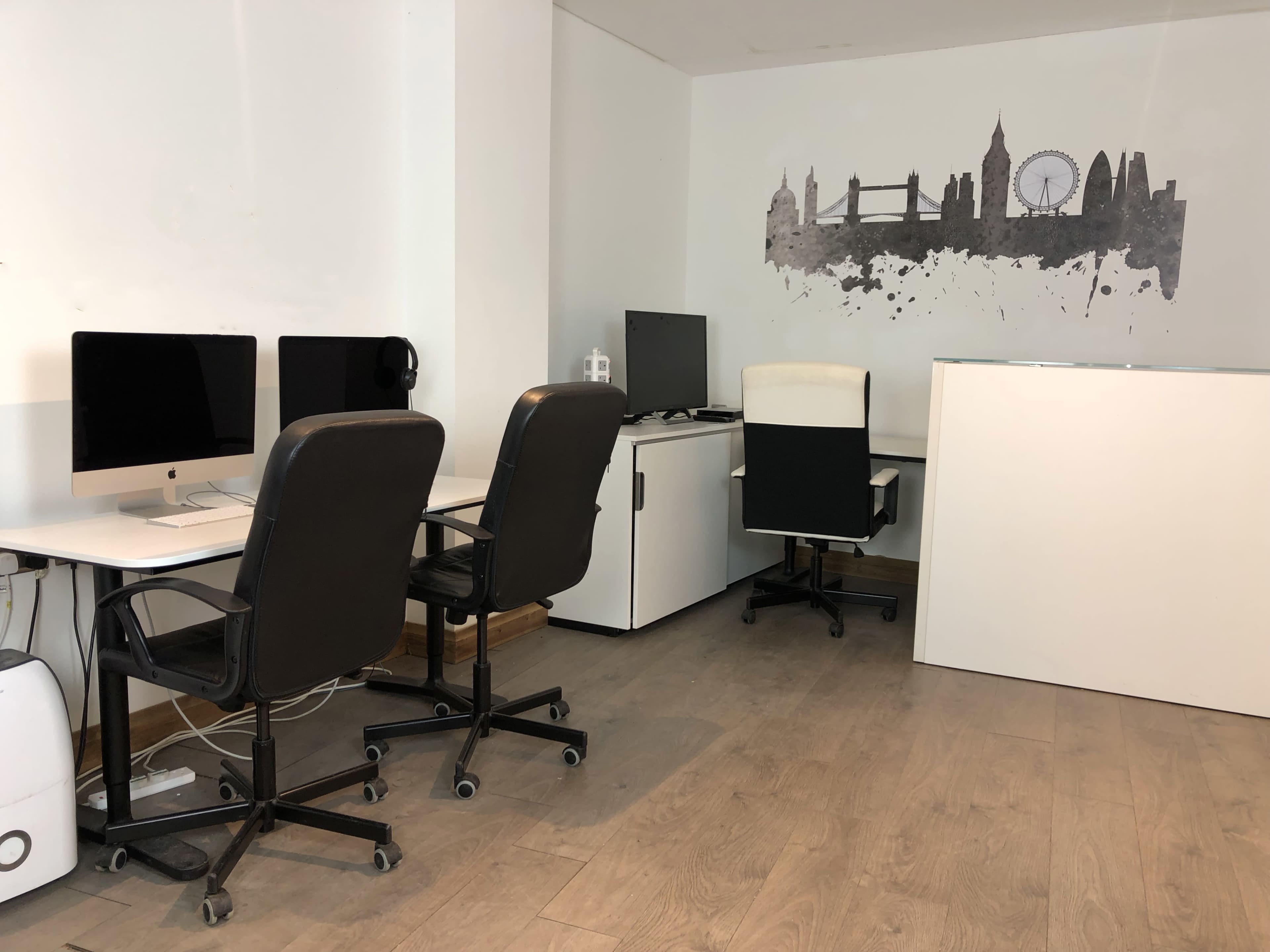 The image shows a modern office space featuring two desks with computers, a chair, and a wall decoration depicting a skyline.