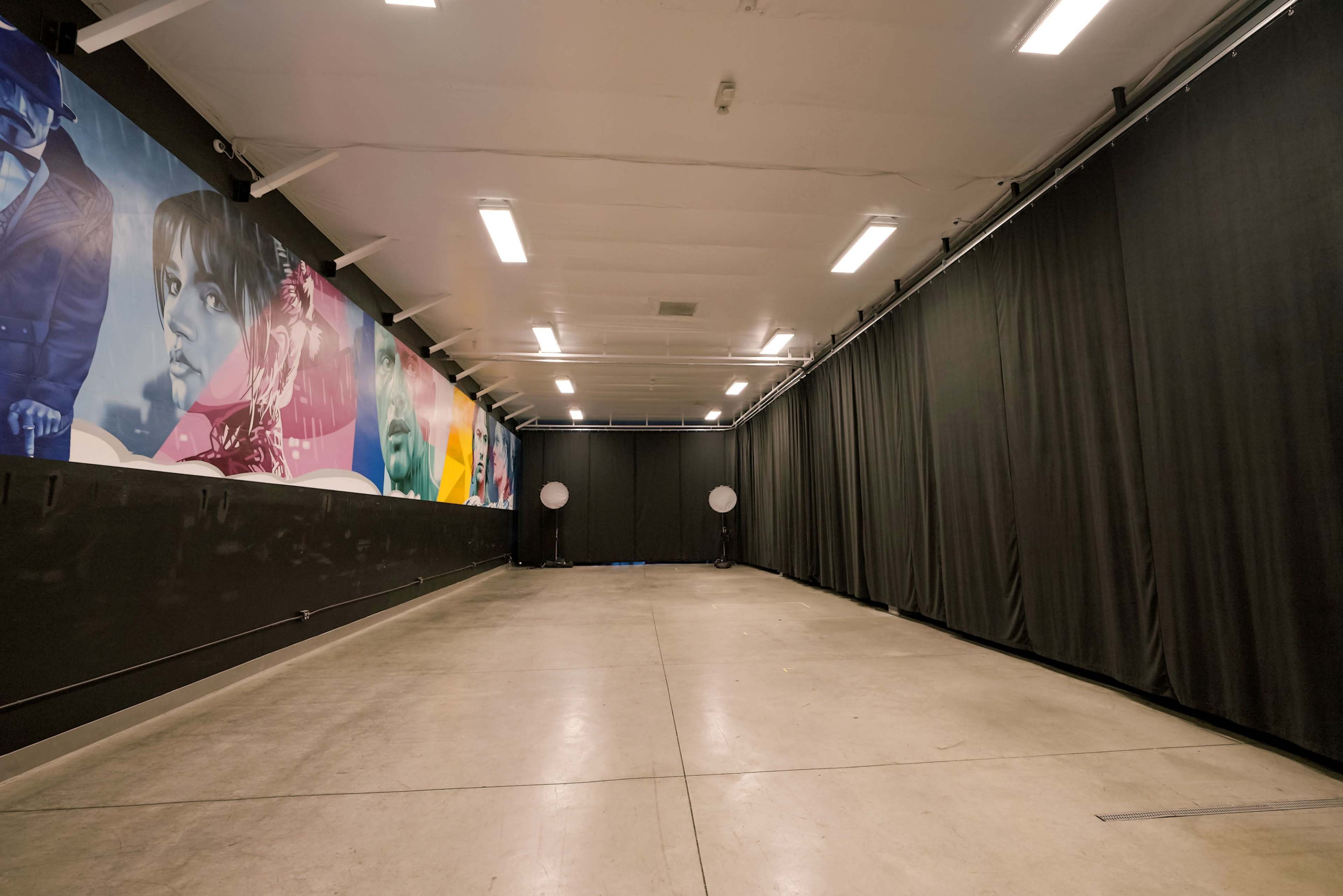 A spacious indoor studio with a concrete floor, black curtains lining the walls, and a colorful mural on one side.