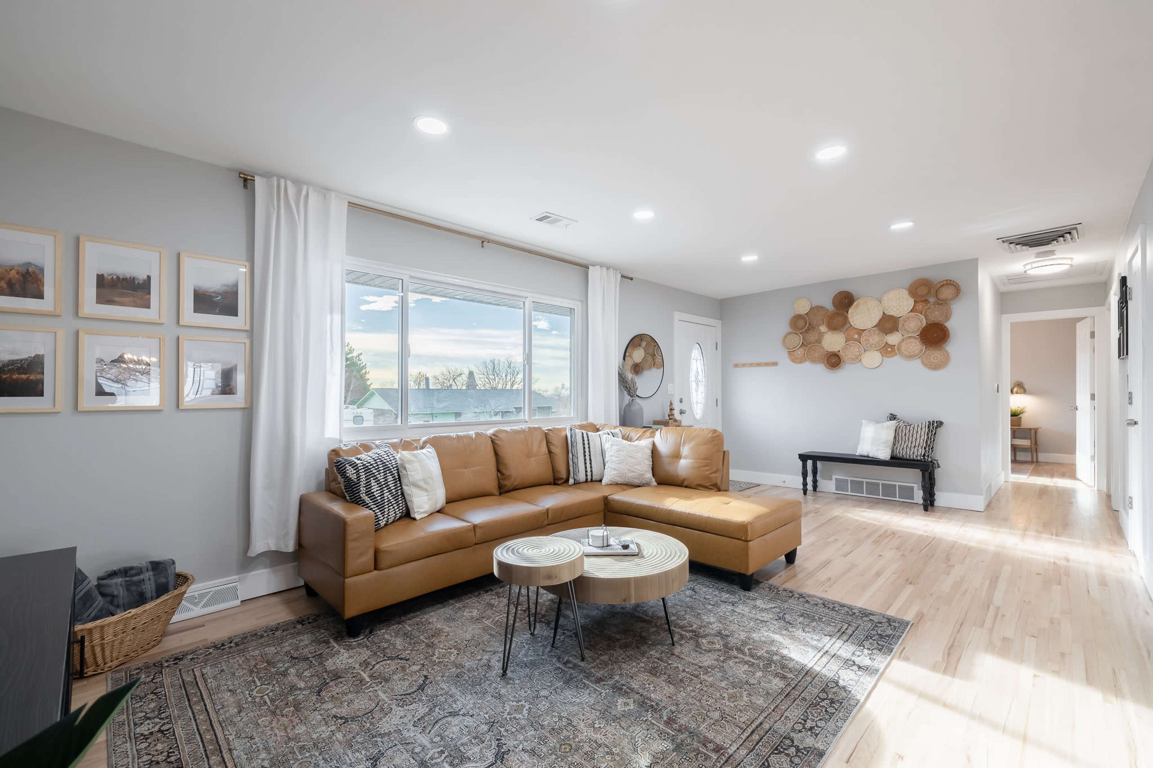 A modern living room features a brown leather sectional sofa, a round coffee table, framed pictures on the walls, and large windows allowing natural light to enter.