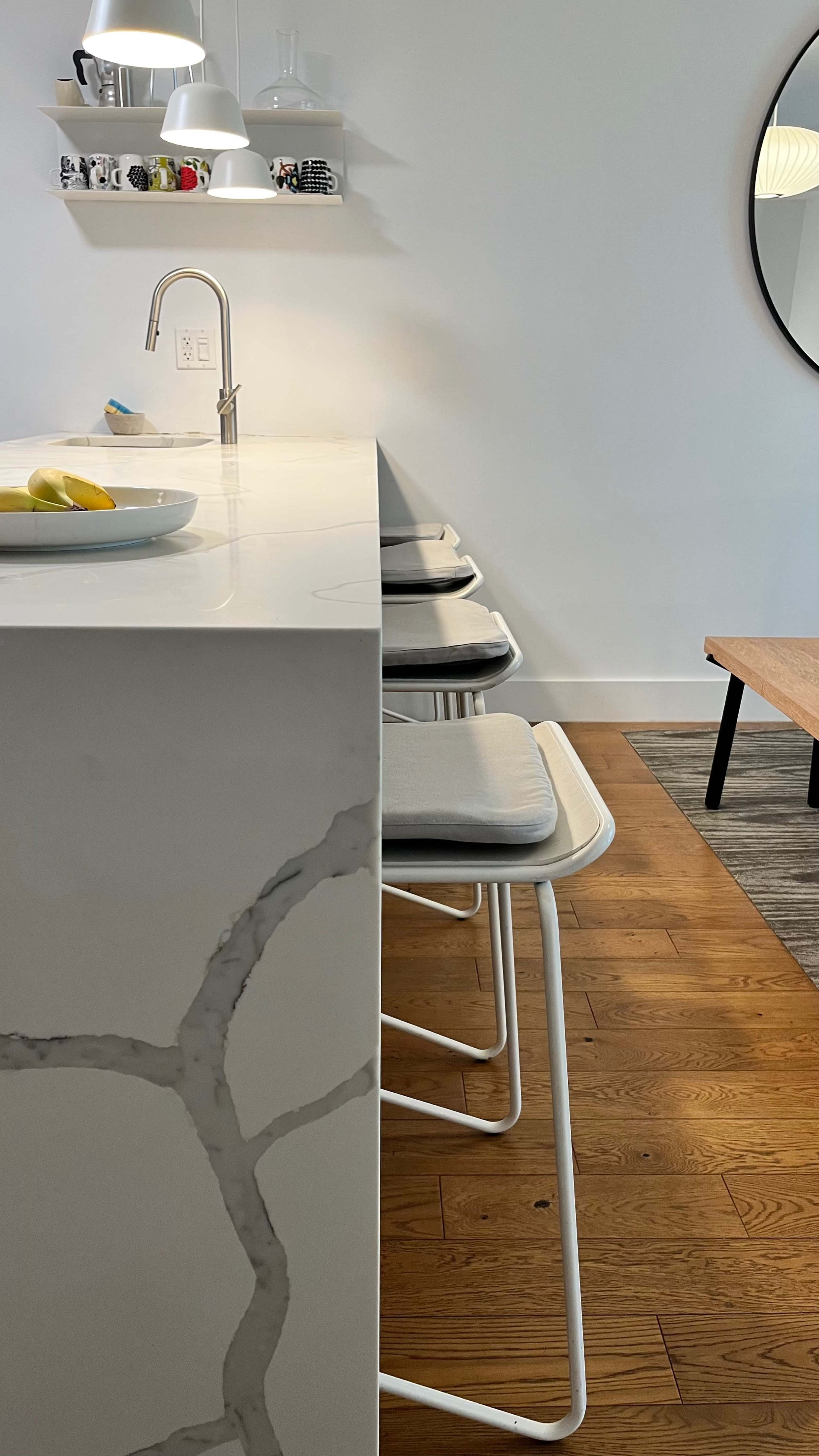 The image shows a modern kitchen bar with three white stools facing a counter, a circular mirror on the wall, and a wooden coffee table nearby.