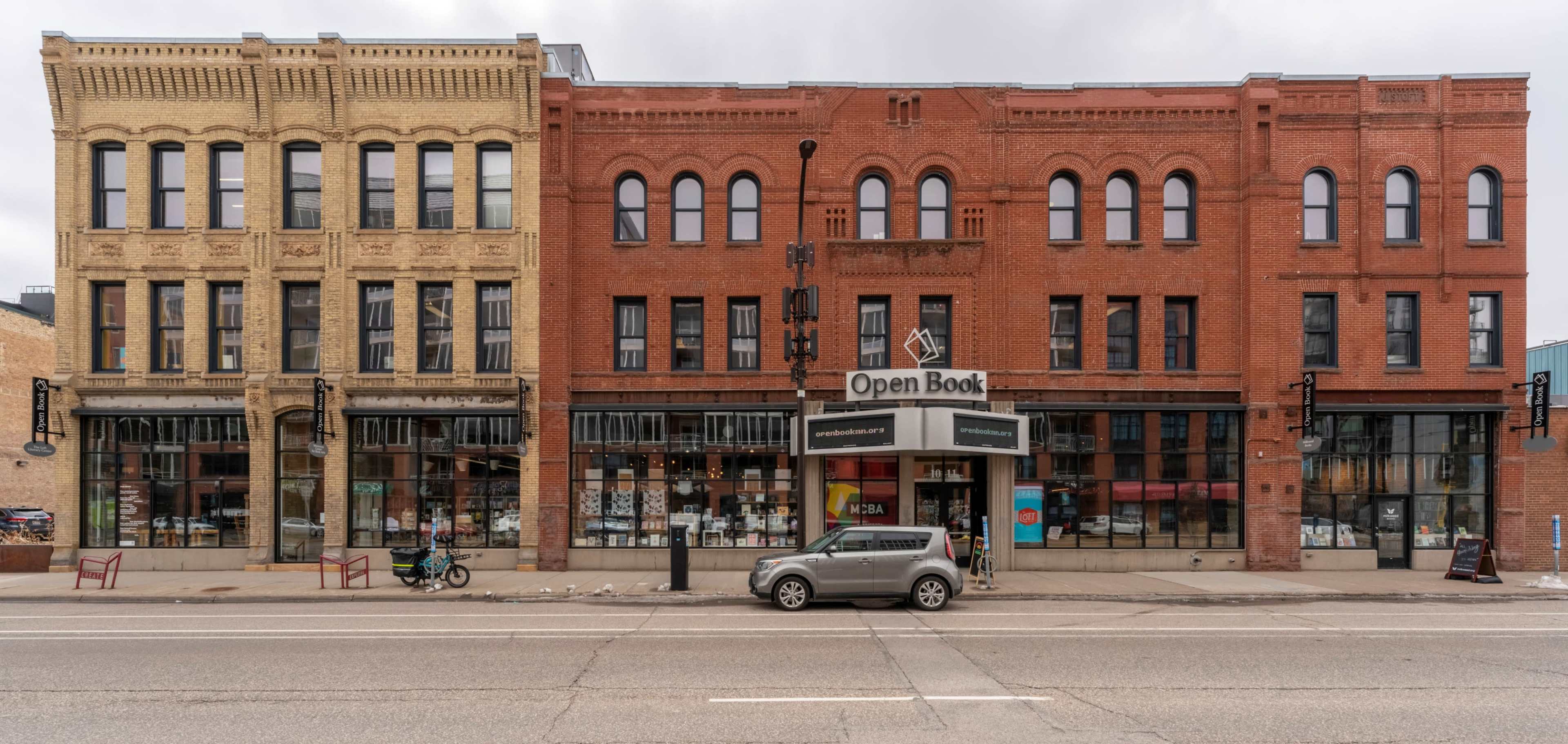 The image shows a street view of a two-story brick building with large display windows featuring a bookstore named "Open Book" at the center, flanked by additional shops.