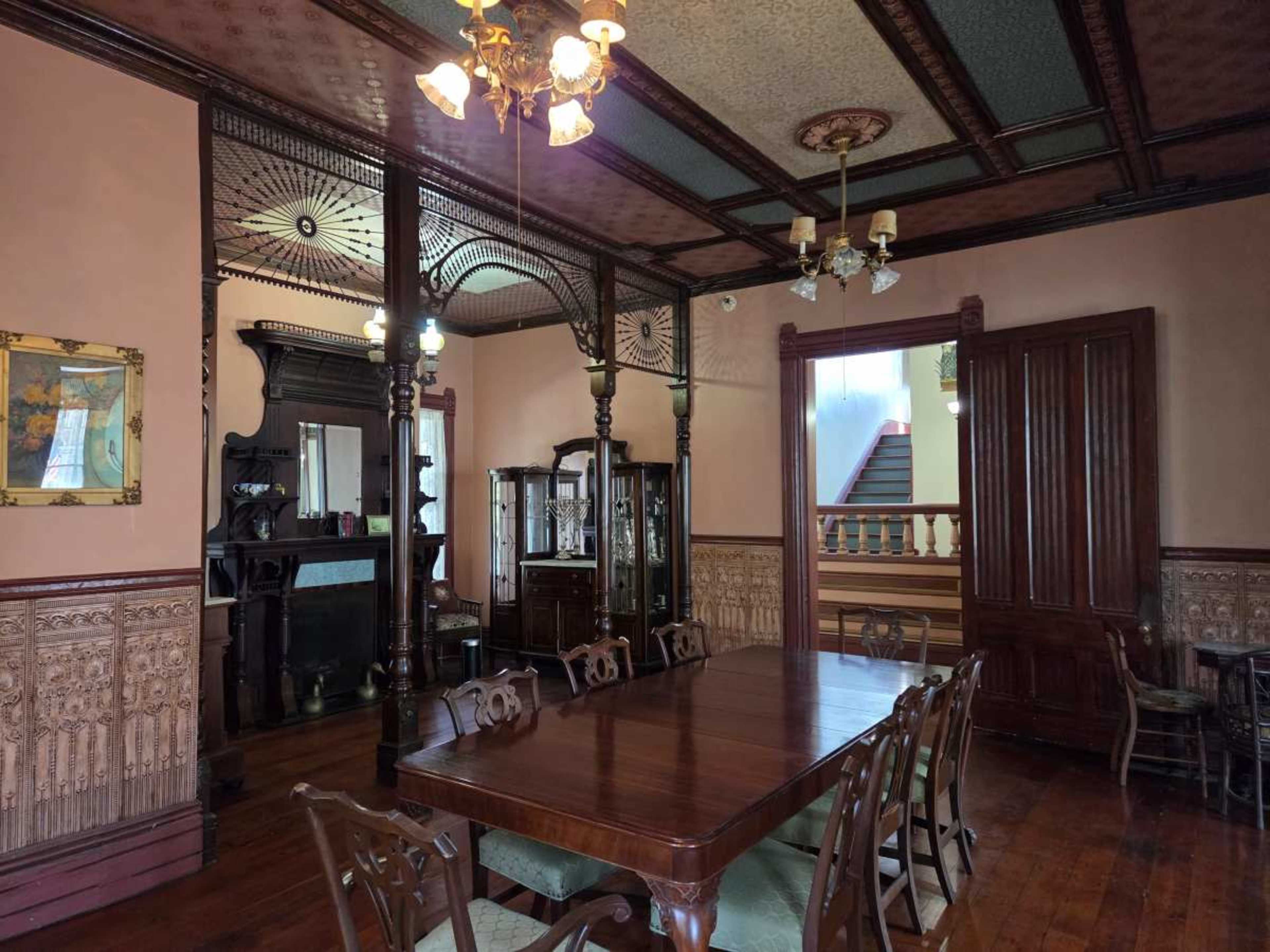 The image shows a vintage dining room with a large wooden table surrounded by chairs, ornate woodwork, and vintage decor, including a mirrored cabinet and a chandelier.