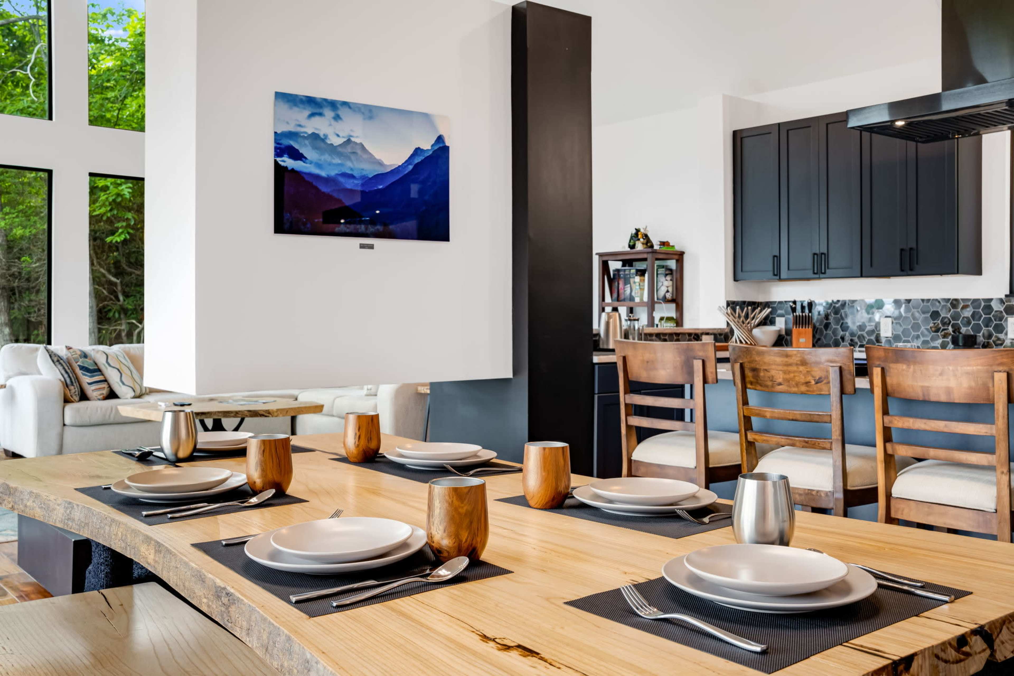 A modern dining area features a wooden table set for four with plates and drinkware, alongside a kitchen with dark cabinets and a wall-mounted television displaying a mountain landscape.