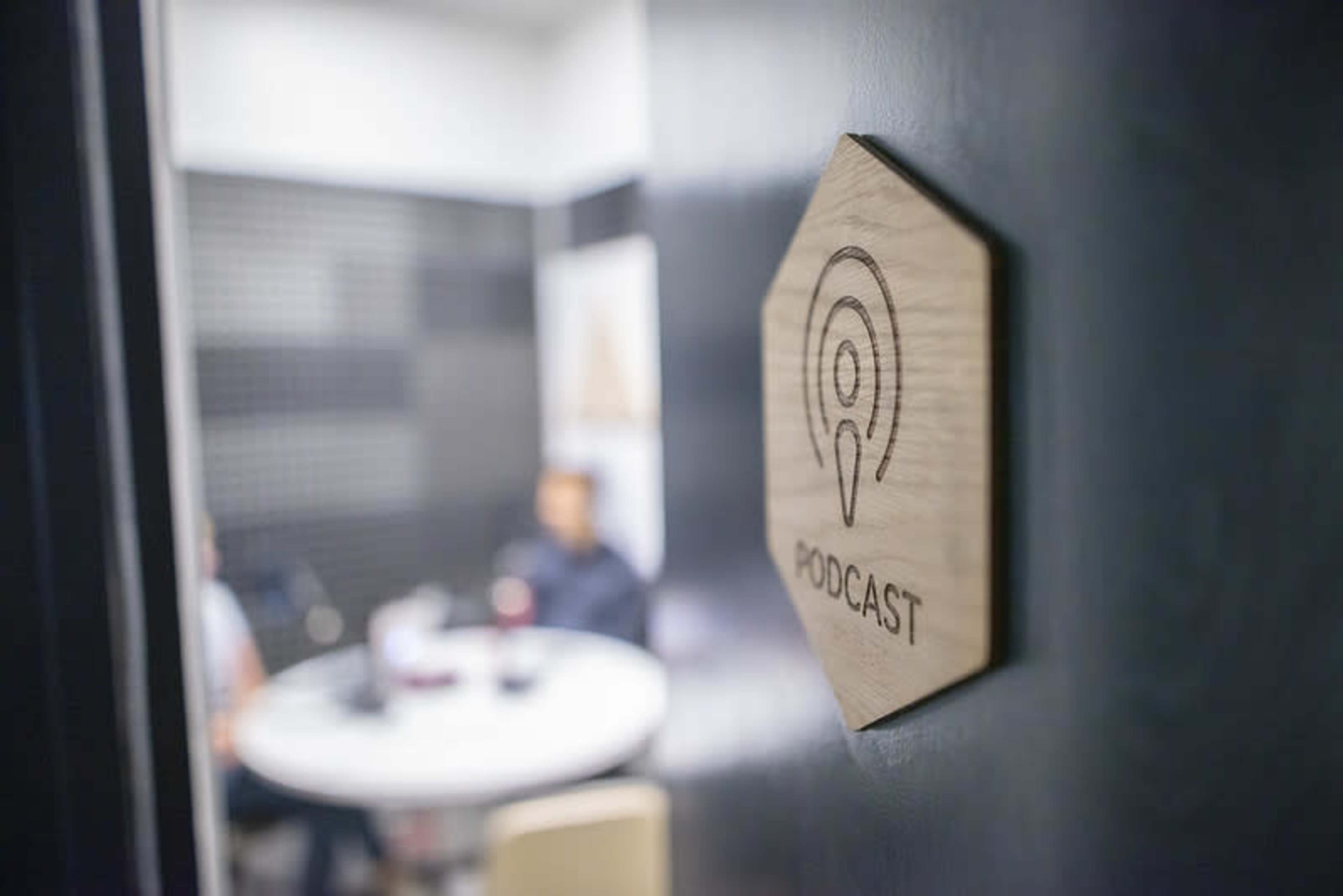A wooden sign labeled "PODCAST" is mounted on a door leading to a small room where two people are seated at a round table.