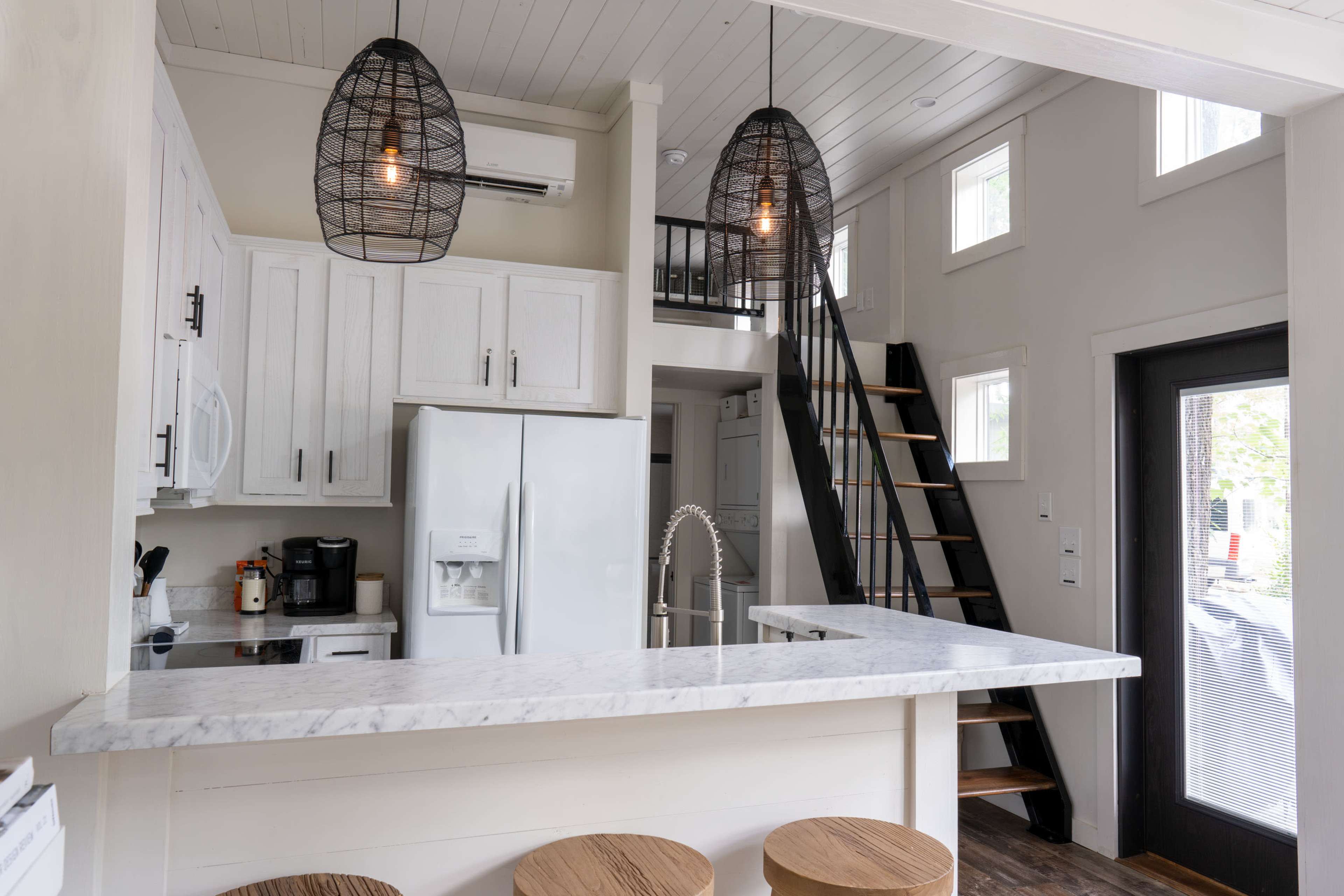 A modern kitchen area with a marble countertop, wooden bar stools, and a staircase leading to an upper level.