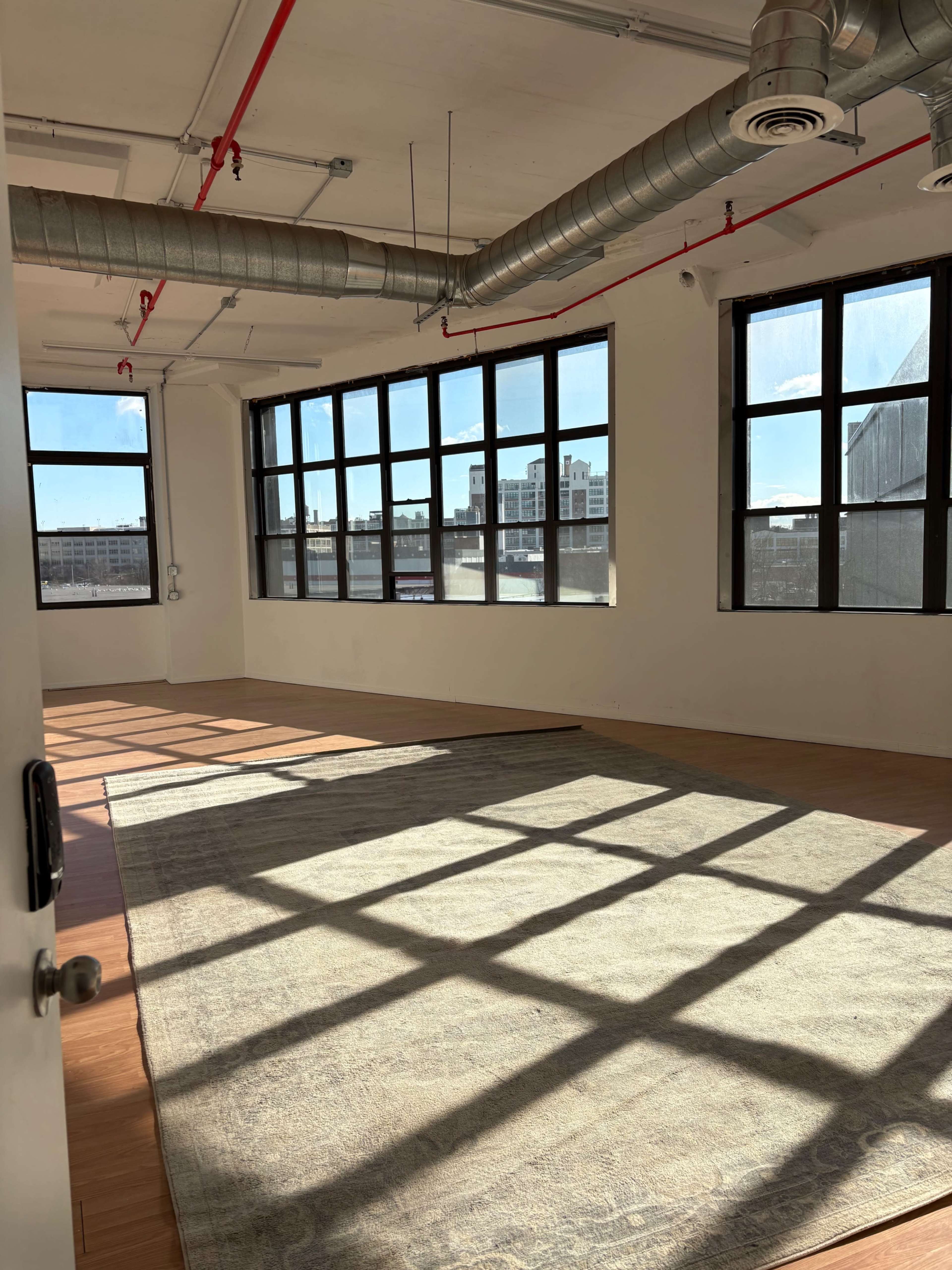 The image shows a spacious, empty room with large windows that allow sunlight to cast shadows on the wooden floor.