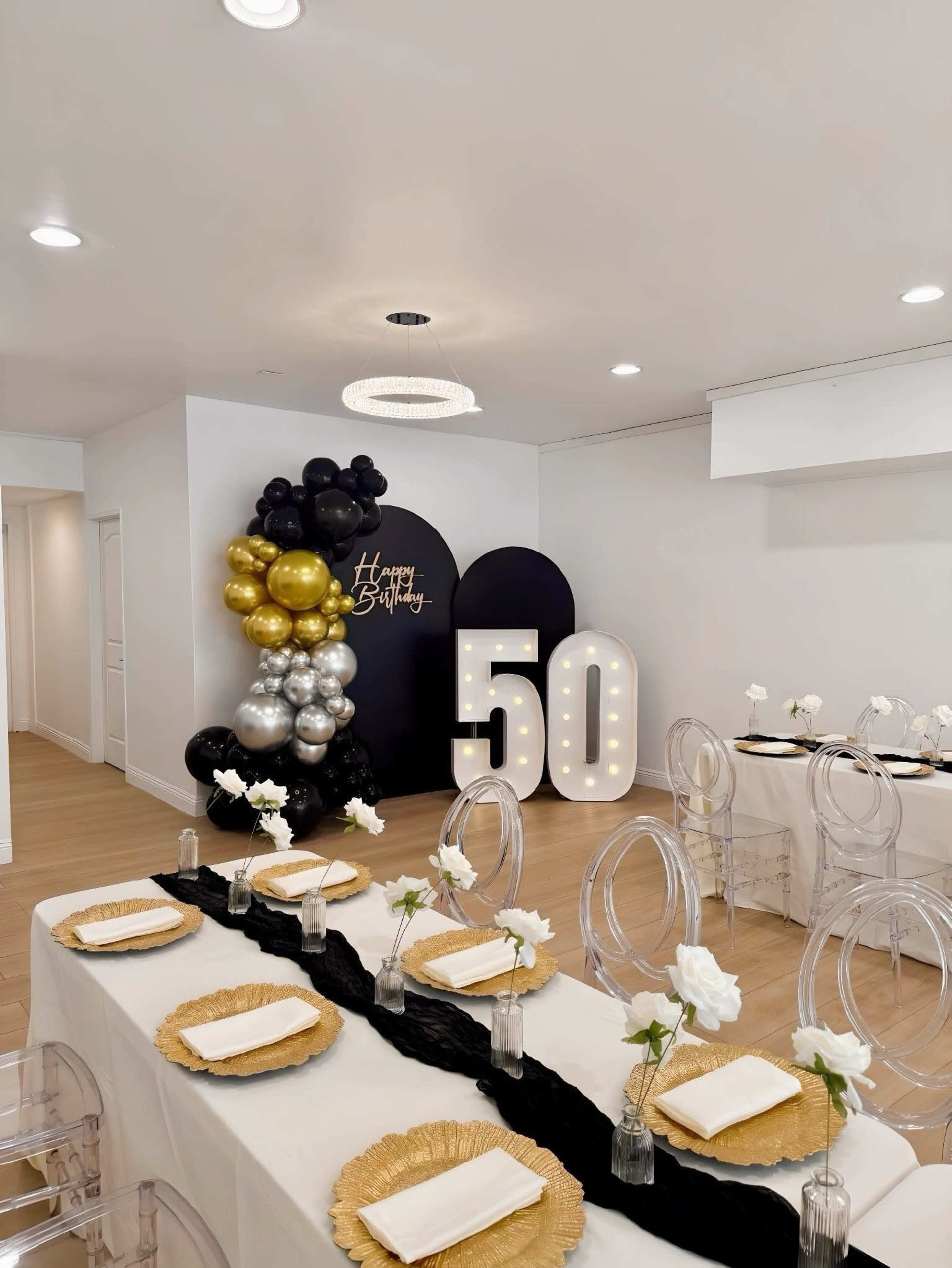 The image shows a dining area decorated for a 50th birthday celebration, featuring a table set with black and gold tableware, clear chairs, and a large illuminated "50" against a wall adorned with balloons.