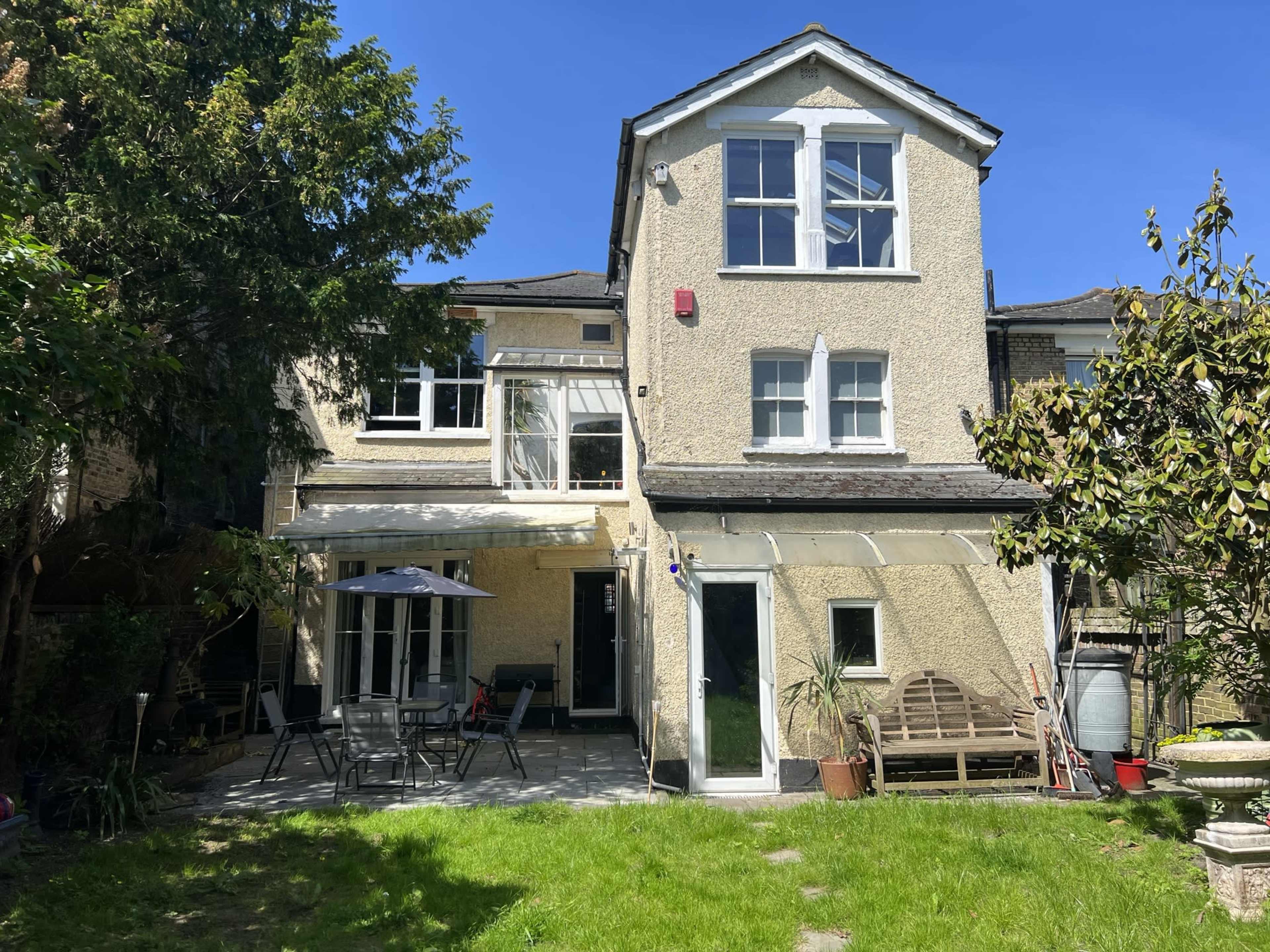 A two-story house with a light-colored exterior, featuring a patio area with outdoor furniture and greenery in the backyard.