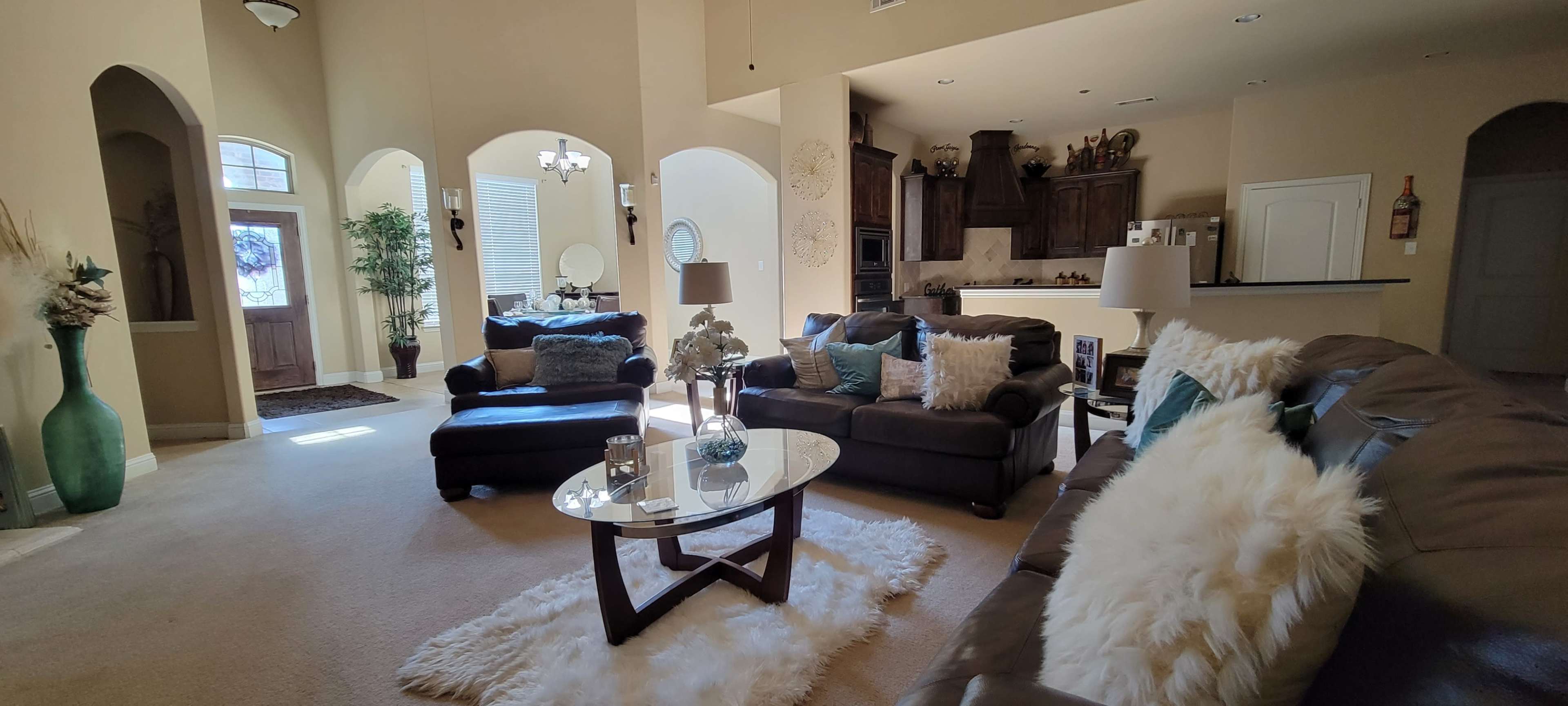 A spacious living room furnished with dark brown sofas, a glass coffee table, and decorative elements, featuring a large window and light-colored walls.