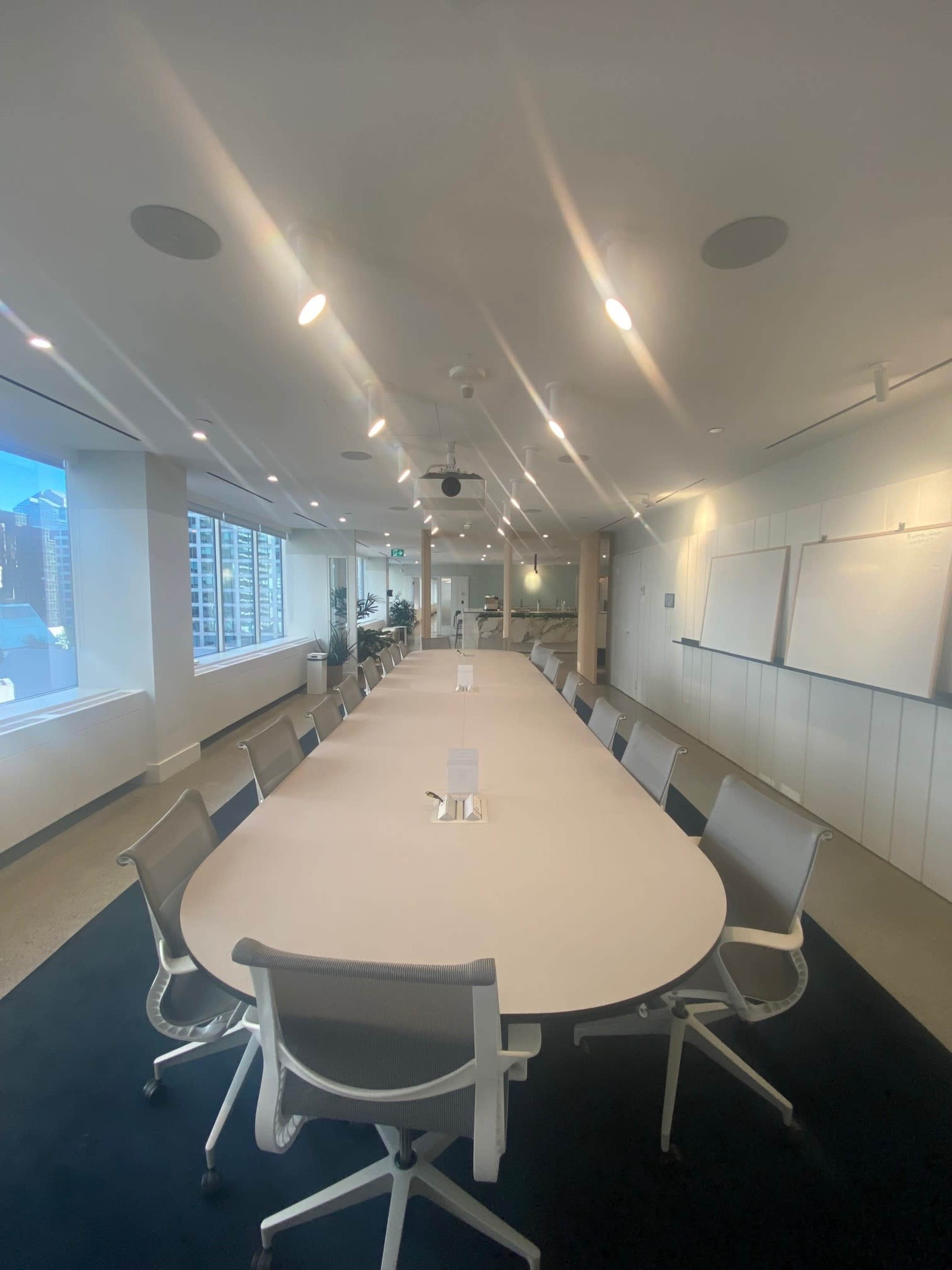 A long, oval conference table surrounded by white chairs is set in a well-lit meeting room with large windows offering a view of a city skyline.