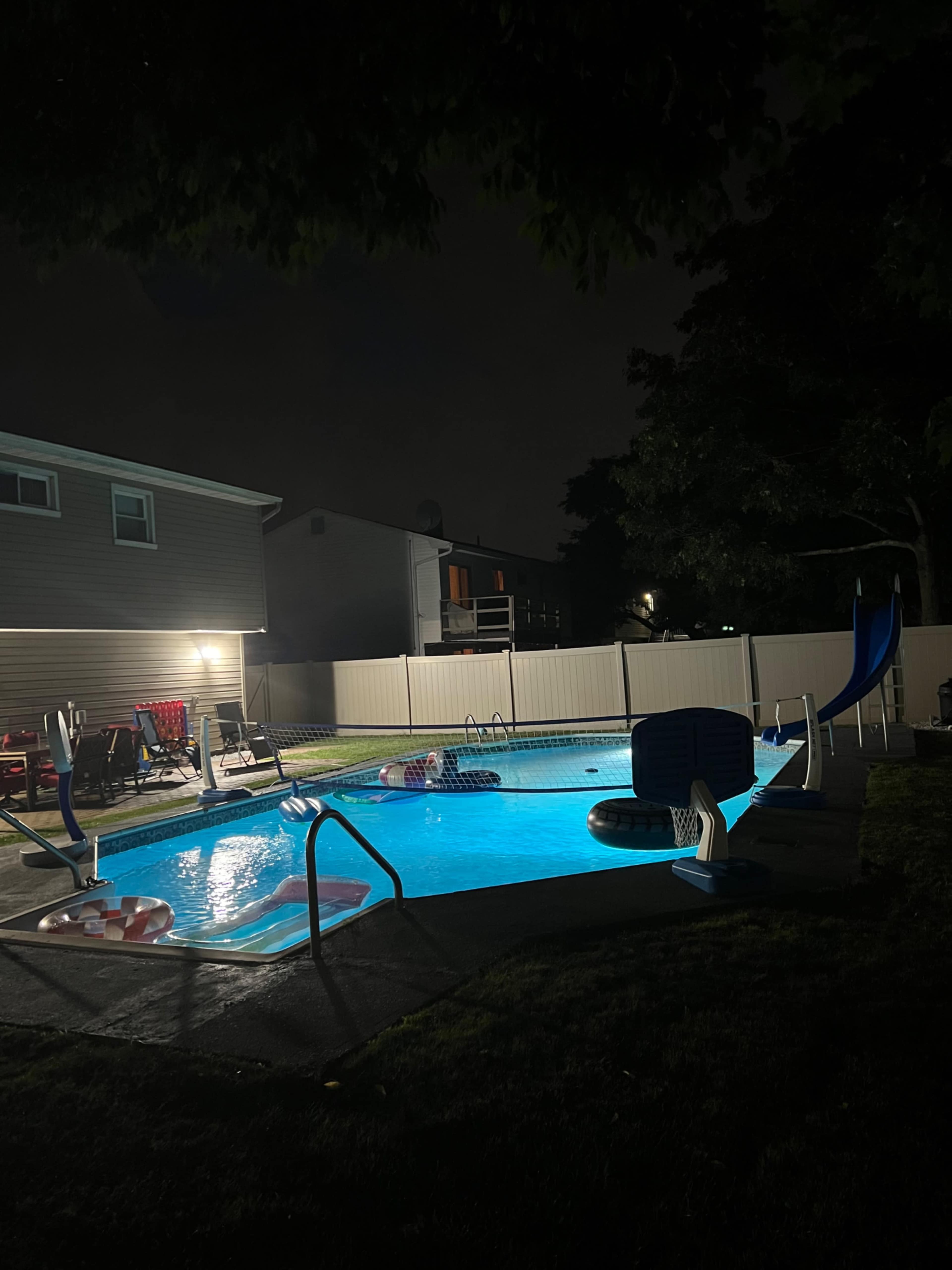 A illuminated swimming pool at night features a slide and lounge chairs in a residential backyard.