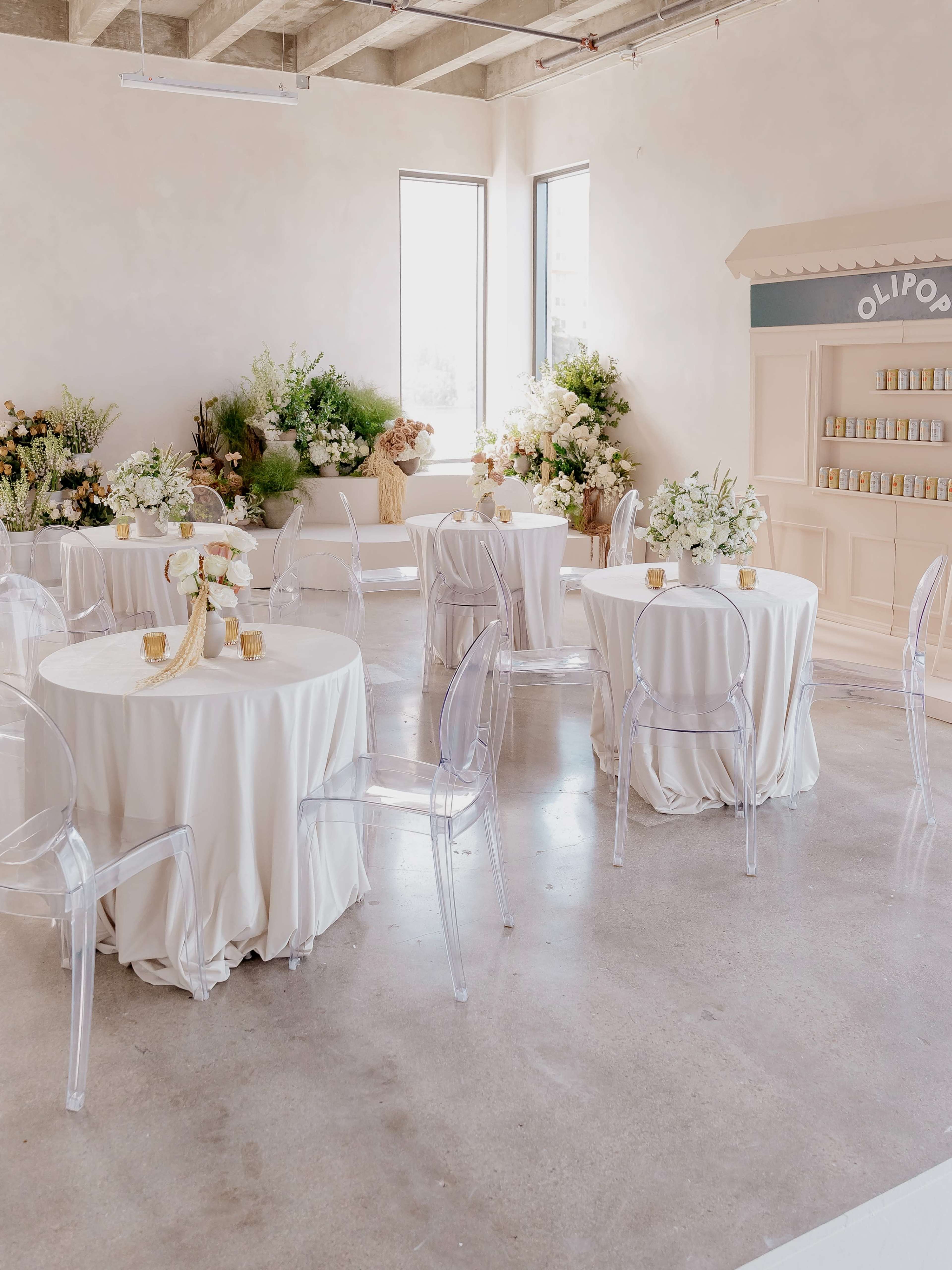 The image shows a well-decorated event space with round tables covered in light-colored tablecloths, transparent chairs, and abundant floral arrangements.