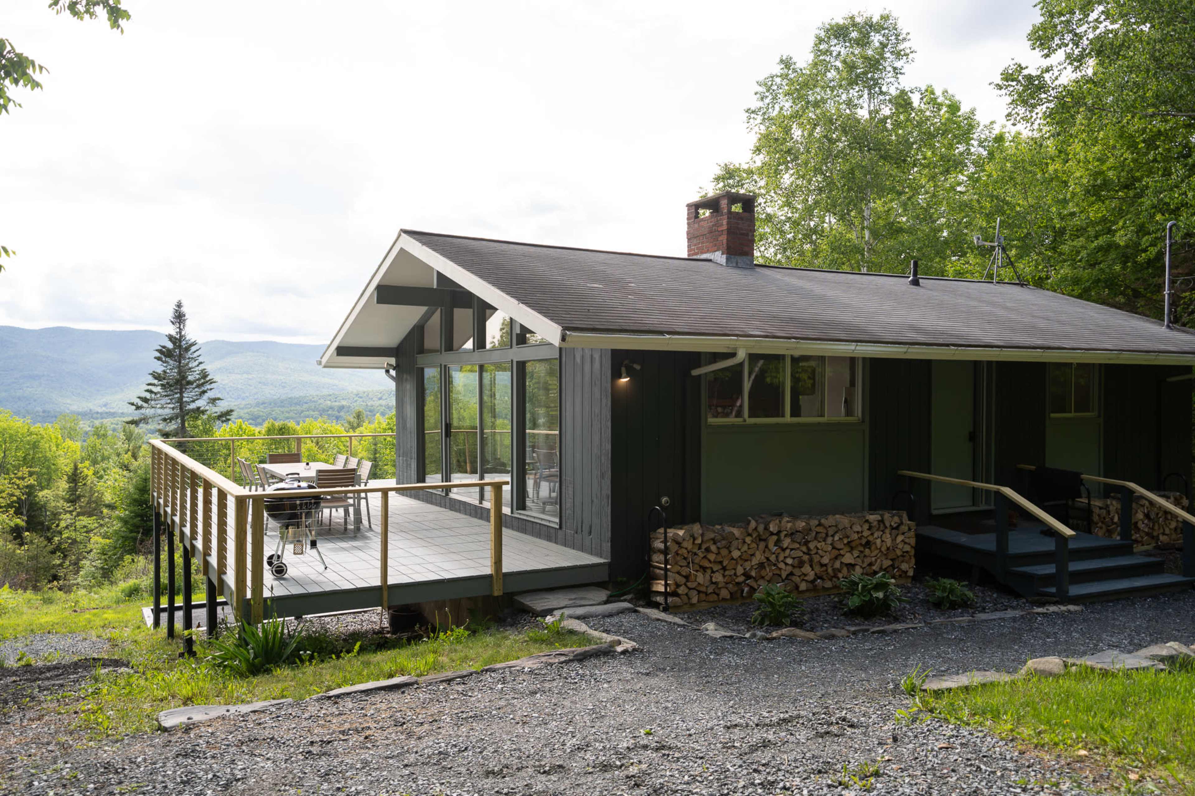 A modern cabin with a deck and stacked firewood overlooks a mountainous landscape.