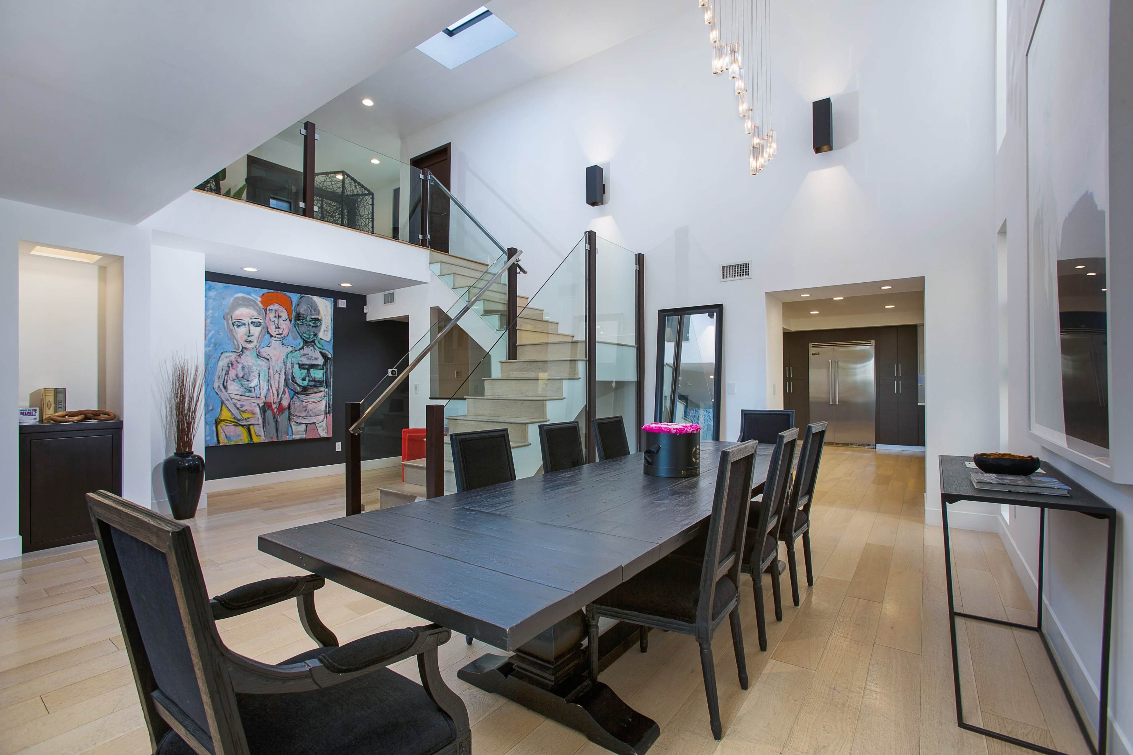 A modern dining area features a large black wooden table surrounded by chairs, with a staircase leading to a mezzanine and artwork displayed on the wall.