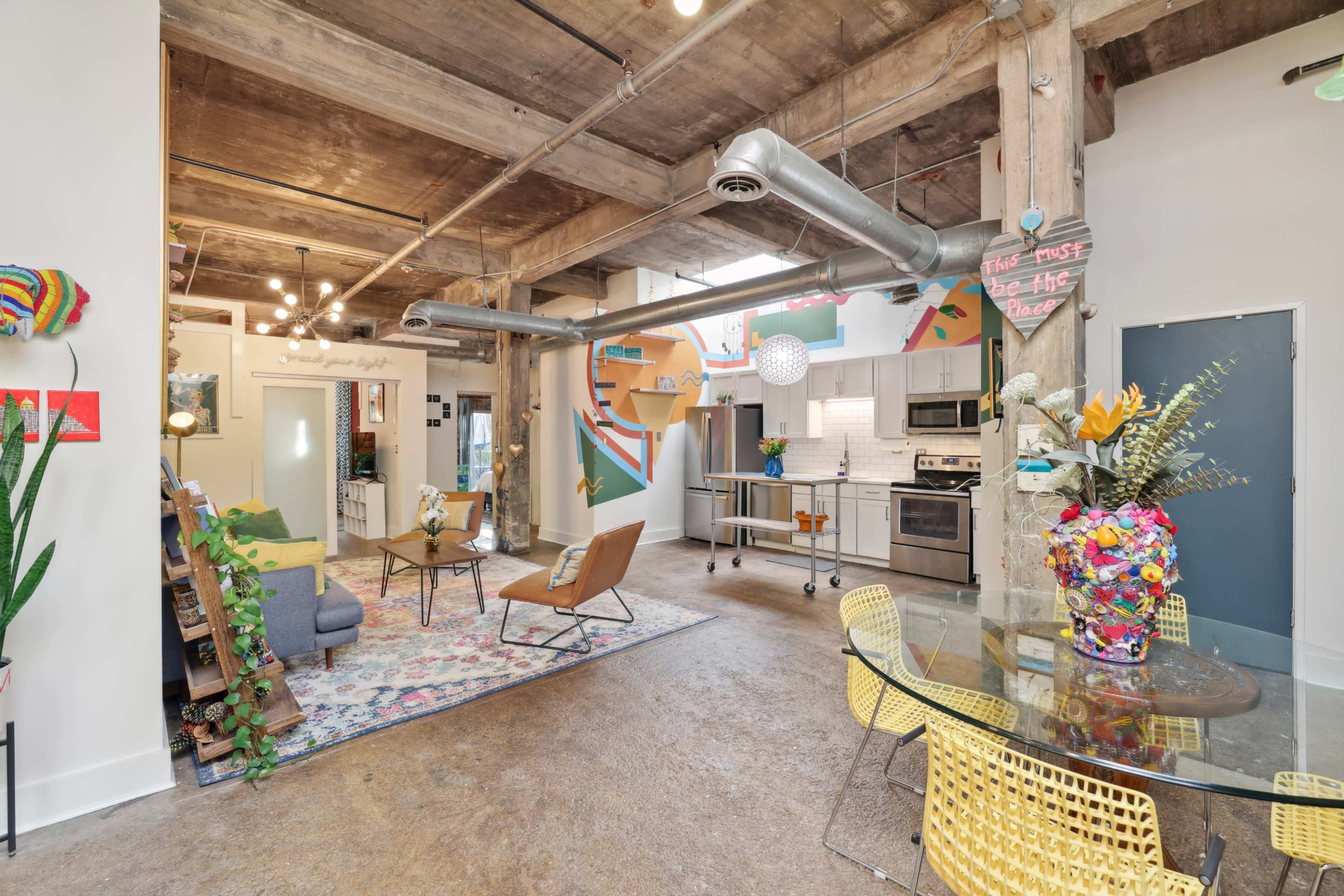 A spacious, urban loft features exposed wooden beams, modern furnishings, and vibrant decorations, including a colorful flower arrangement on a dining table.