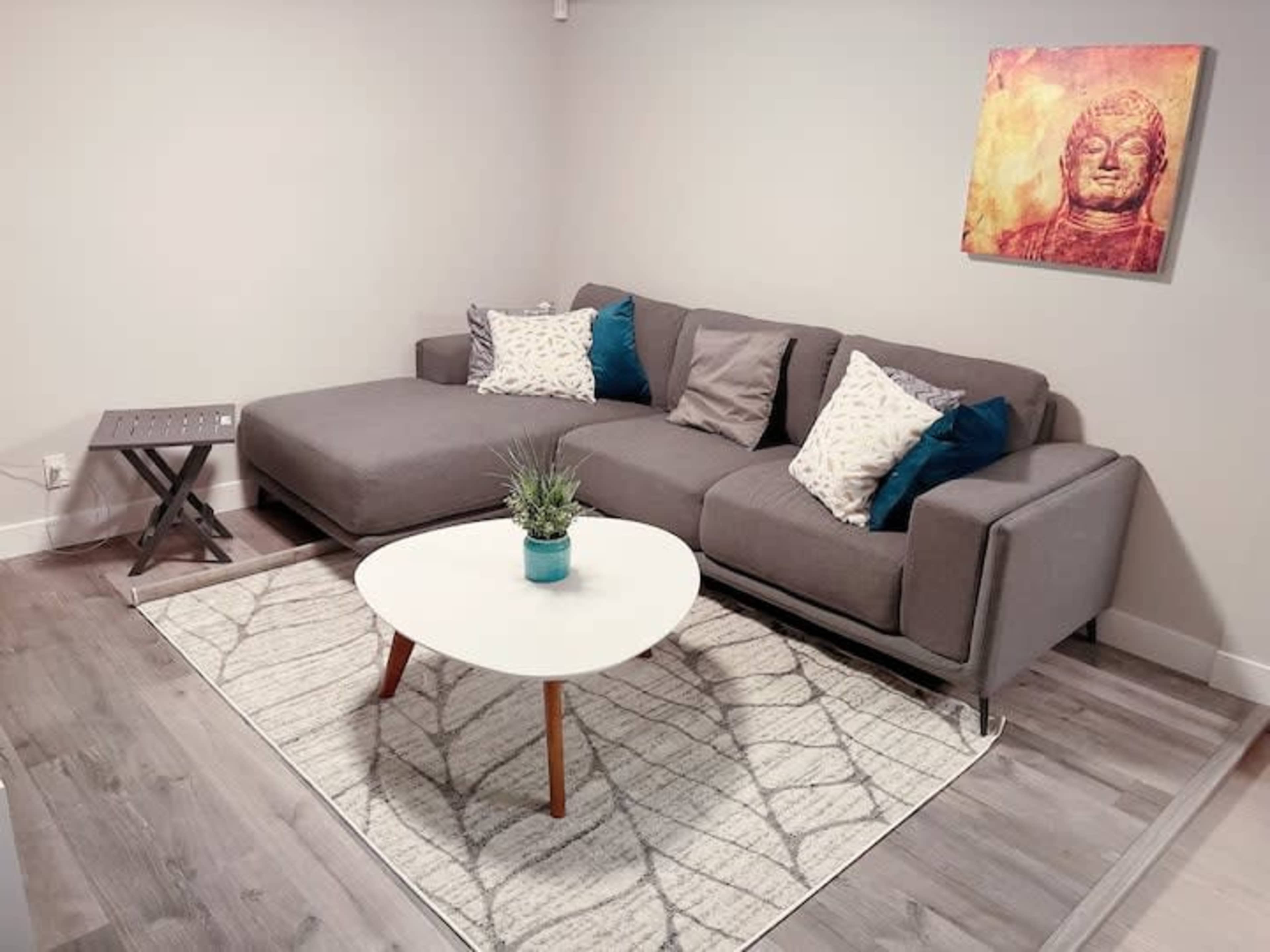 A gray sectional couch with patterned pillows is positioned next to a small side table, facing a round coffee table with a potted plant, all placed on a decorative rug in a bright room.
