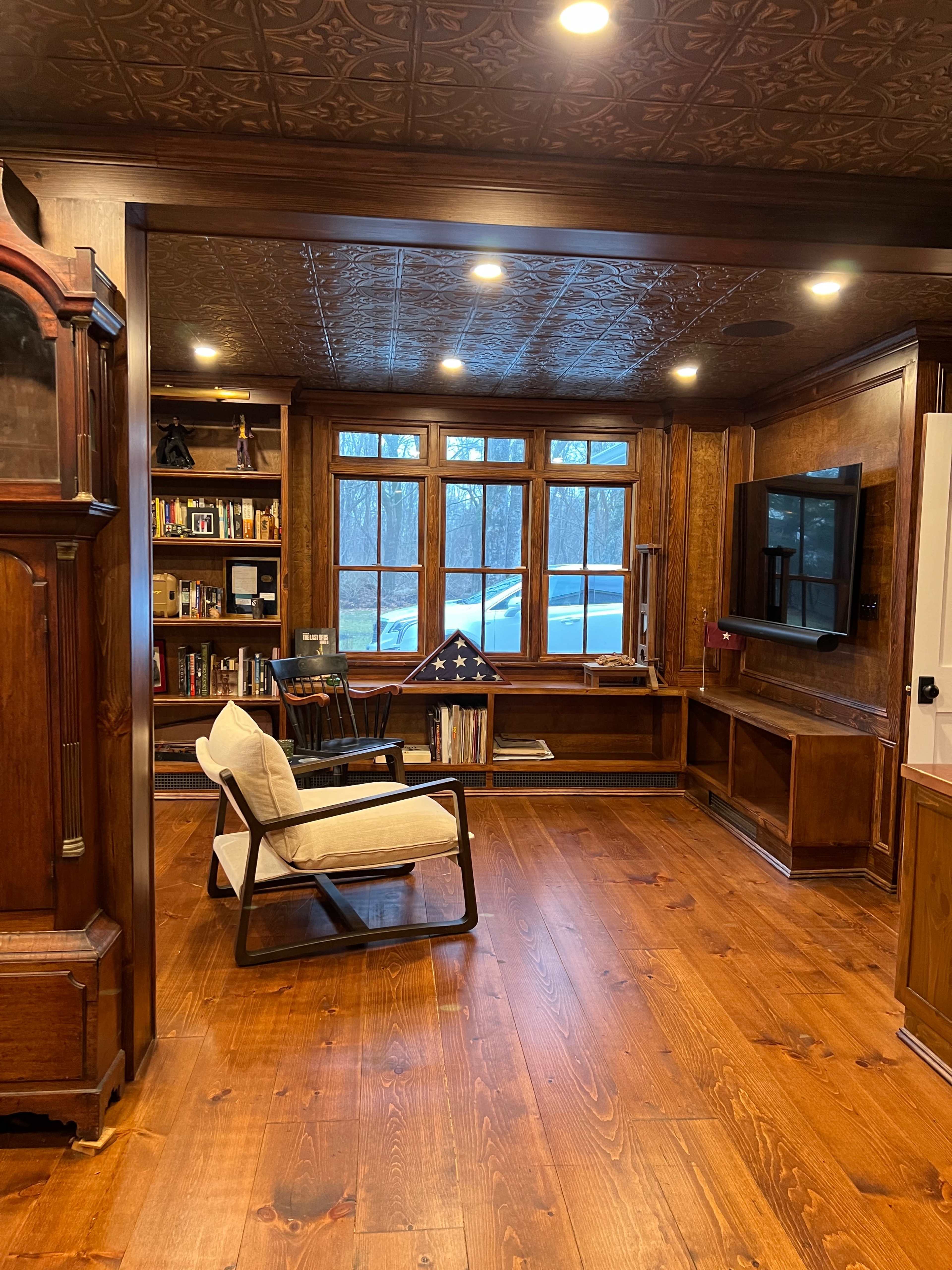 A cozy room with wooden walls, a large window, bookshelves filled with books, a chair, and a television mounted on the wall.