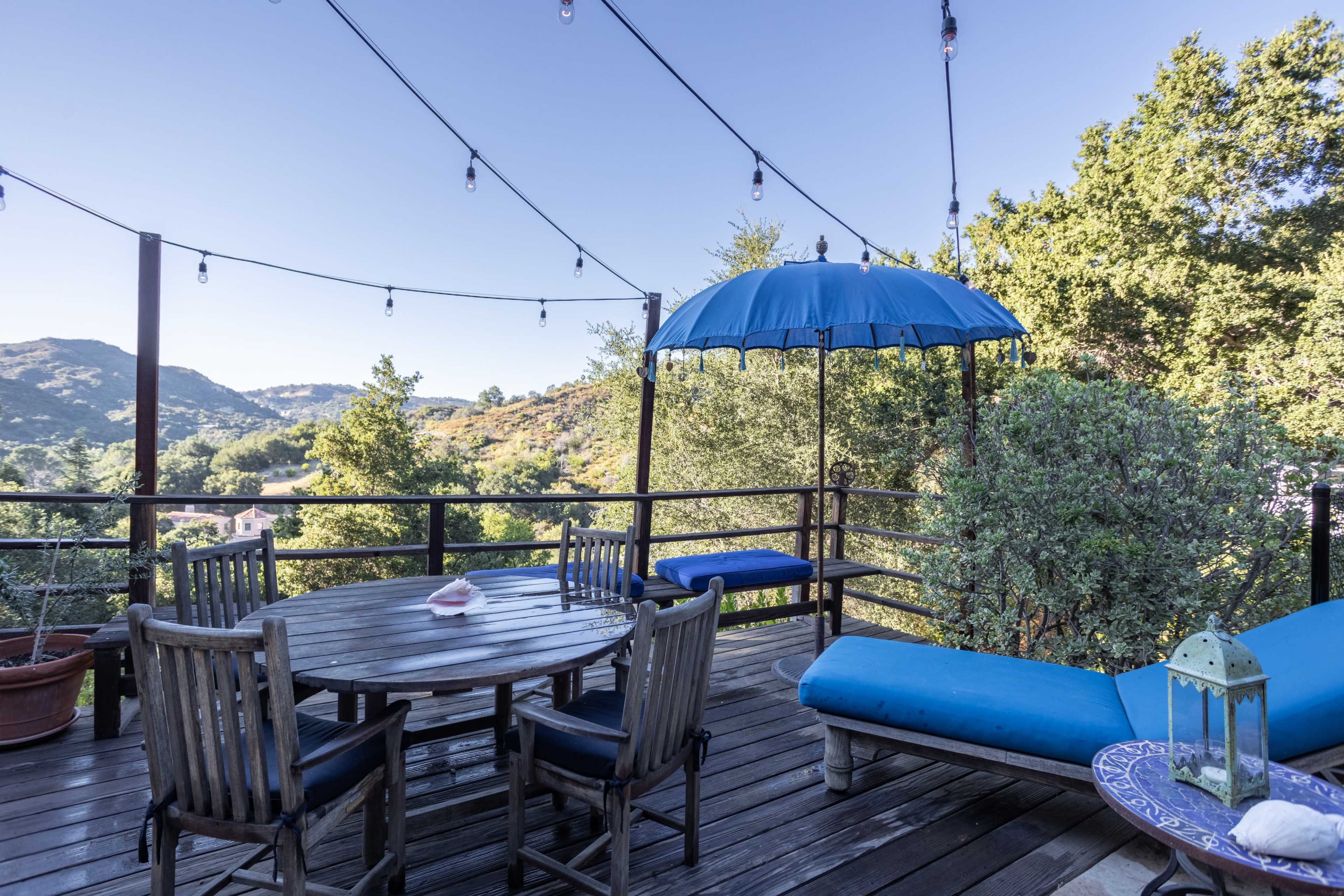 A wooden deck features a round dining table, a lounge chair, and an umbrella, surrounded by trees and hills under a clear sky with string lights overhead.