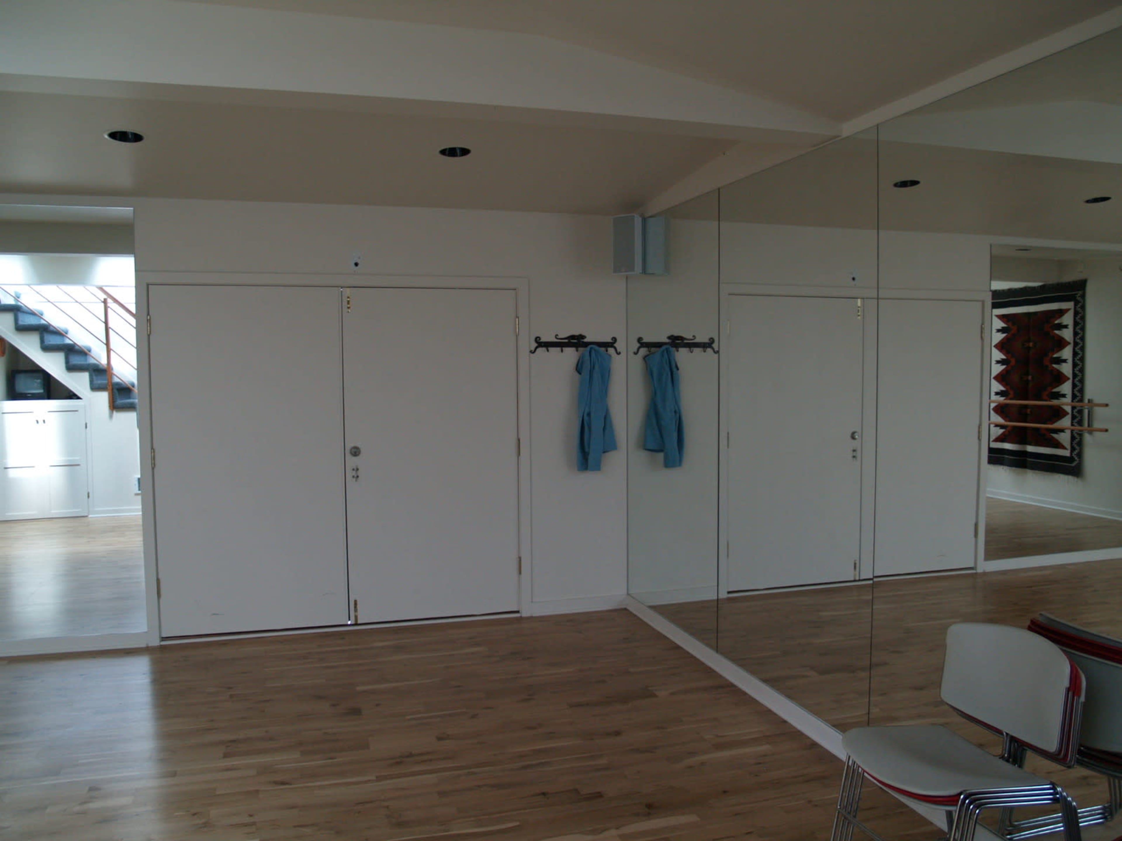 A spacious room with wooden flooring, a large mirror on one wall, and two blue garments hanging on hooks near a closed door.