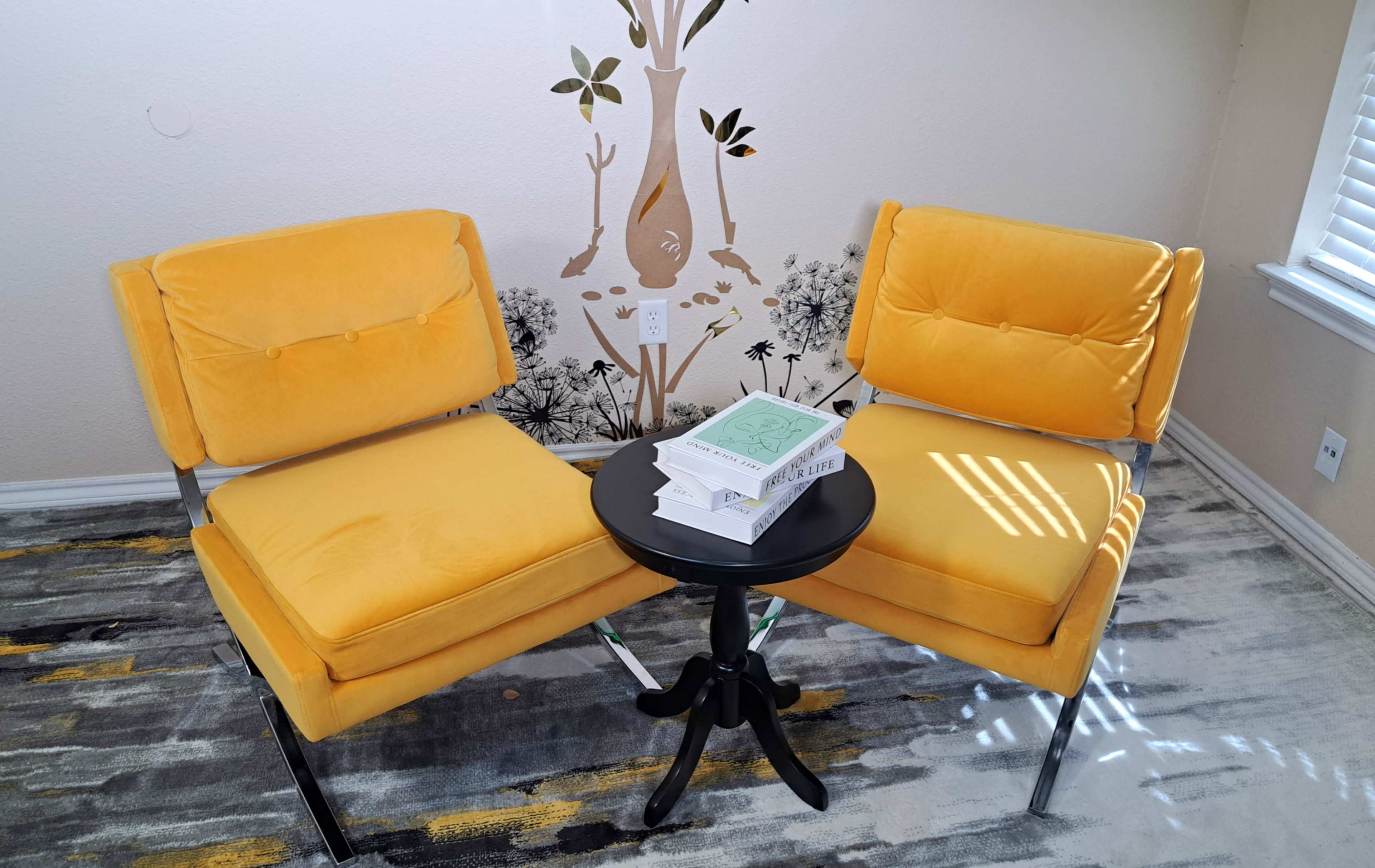 The image shows two yellow chairs facing each other around a black circular side table, with a stack of books on top, set against a wall featuring a decorative design.