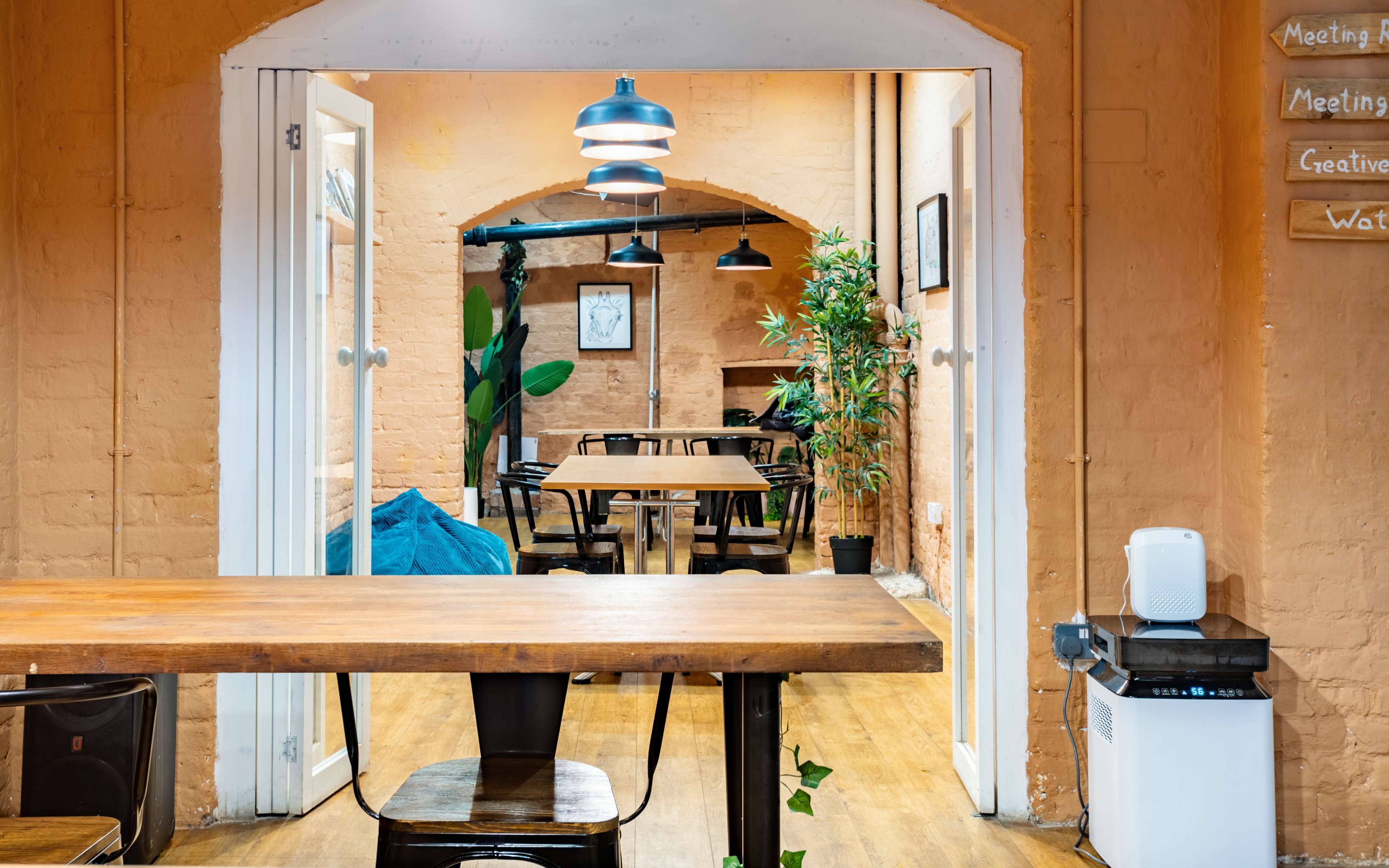 A spacious, well-lit communal area with wooden tables, black chairs, green plants, and archways leading to additional seating spaces.