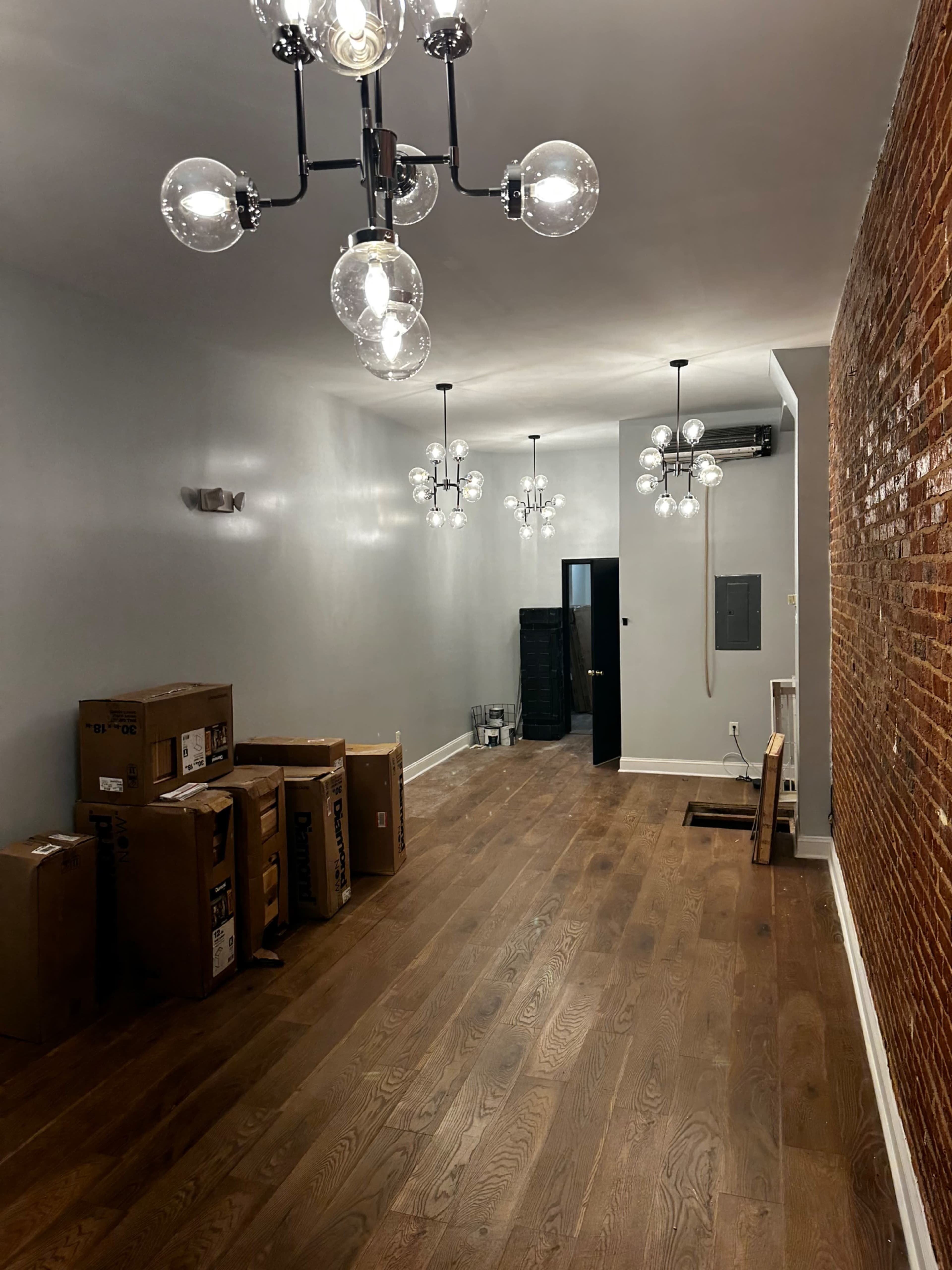 The image shows a narrow, empty hallway with exposed brick walls, wooden flooring, and several boxes stacked along one side, illuminated by multiple modern light fixtures.