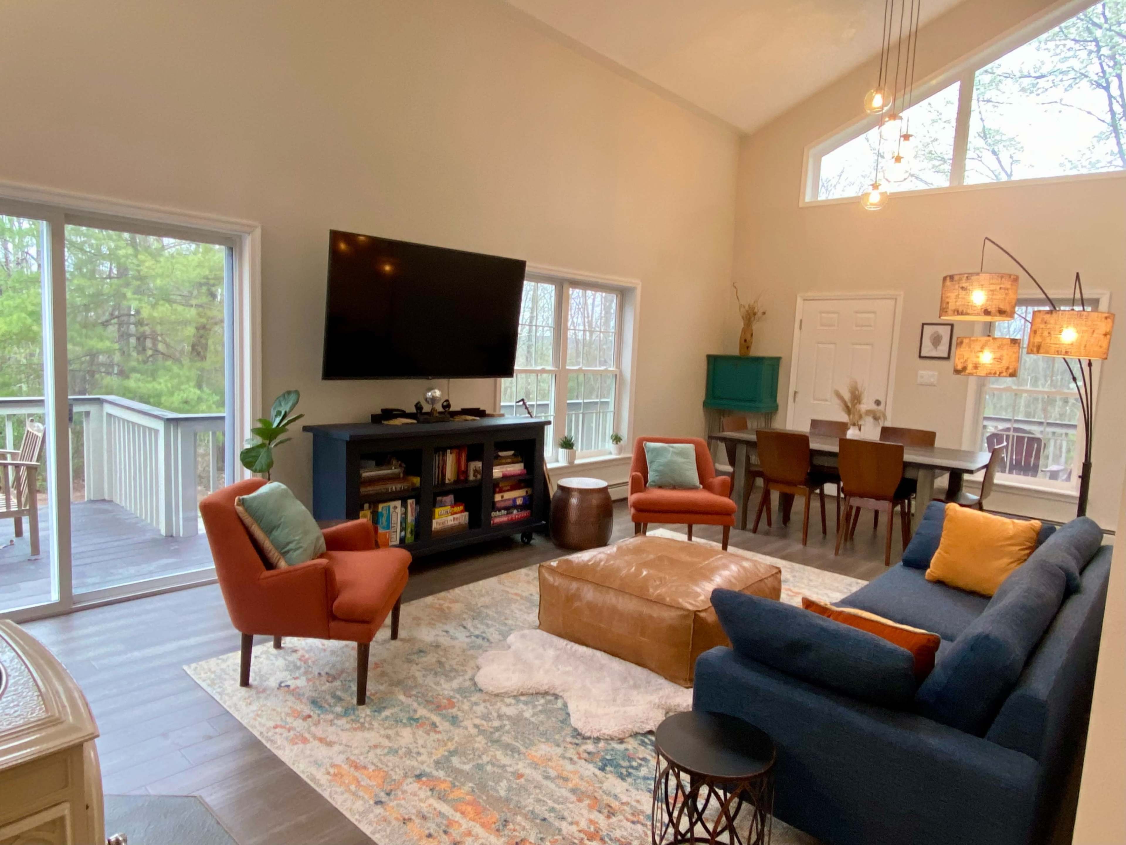 The image shows a modern living room with a blue sofa, a wooden coffee table, a large television mounted on the wall, and a dining area in the background, featuring a table and chairs.