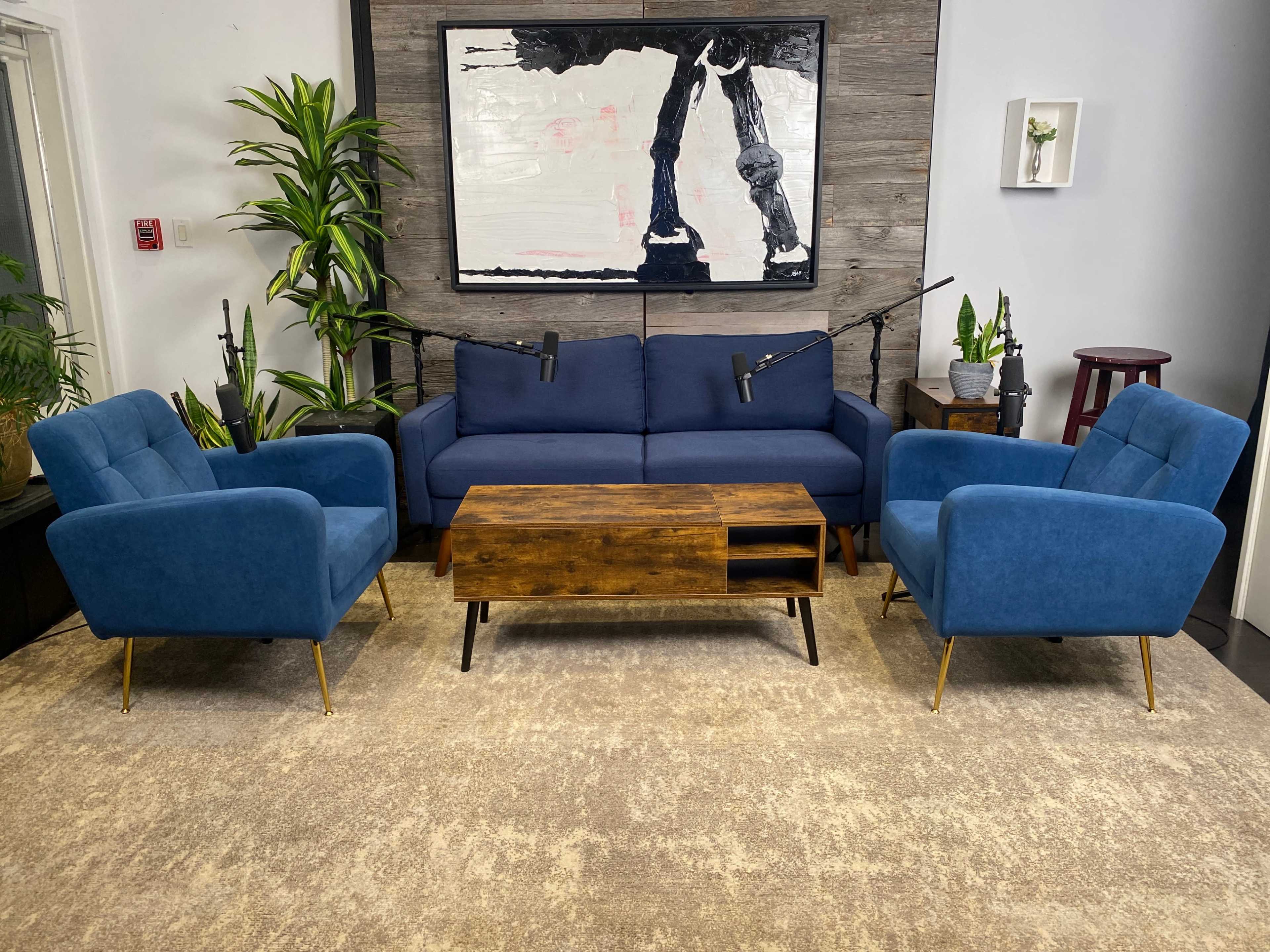 A modern, minimalist living room setup with a blue sofa, two matching armchairs, a wooden coffee table, and decorative plants against a neutral background.