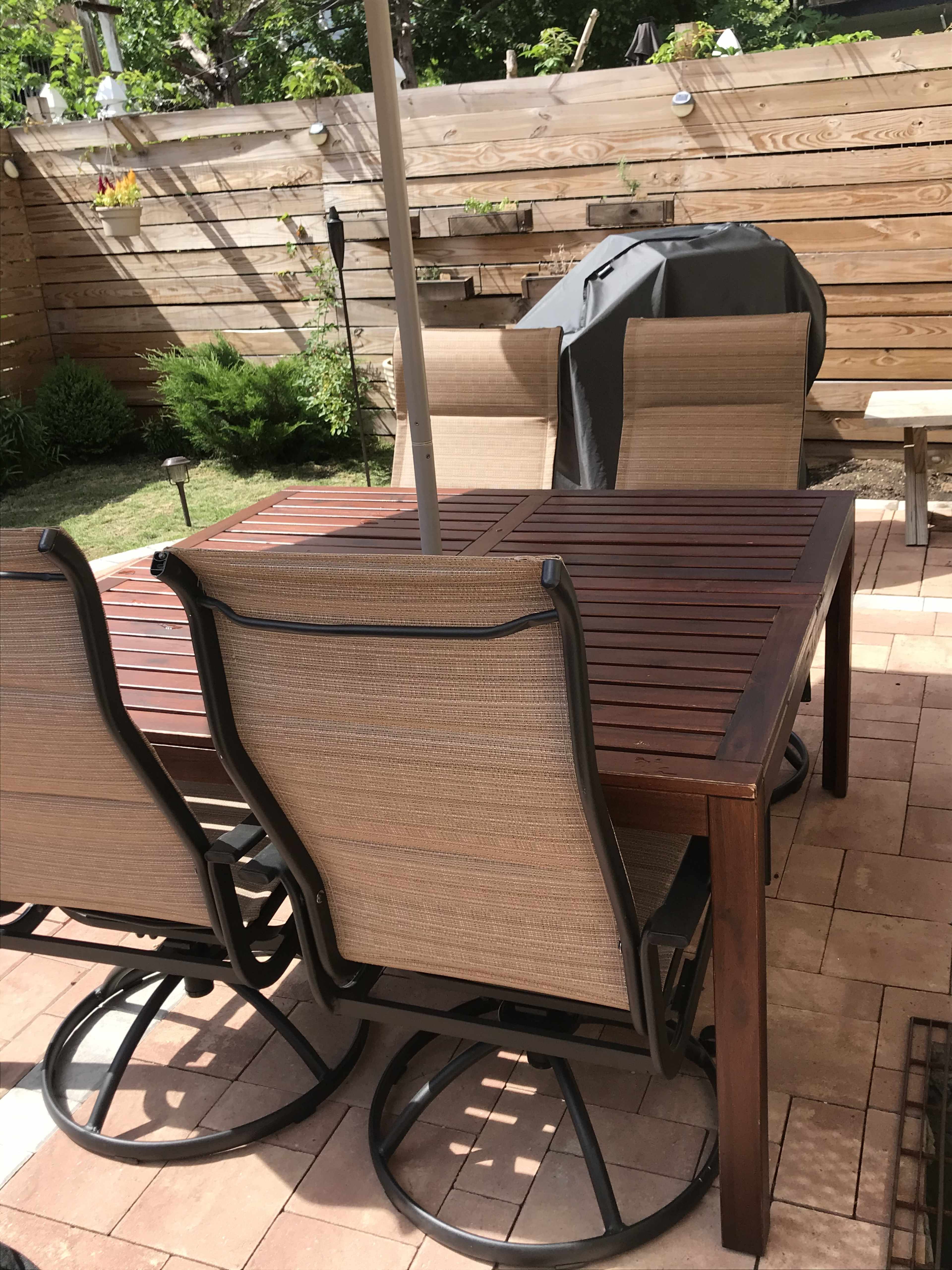 A wooden dining table with four swivel chairs sits on a patio, surrounded by greenery and a grill under an umbrella.