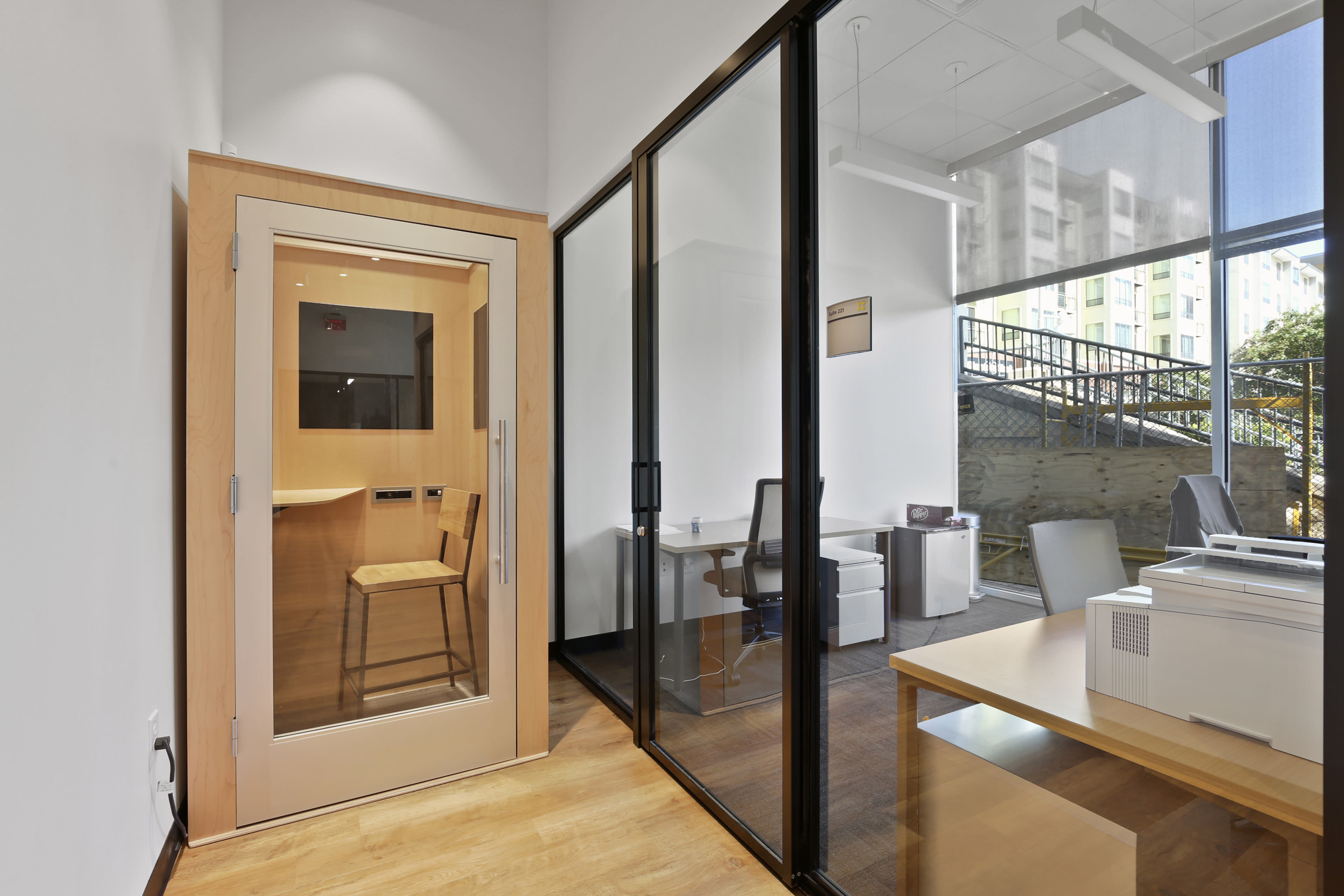 The image shows a modern office space featuring a glass-walled meeting room, a wooden enclosed area with a chair, and a workspace with a printer and desk.