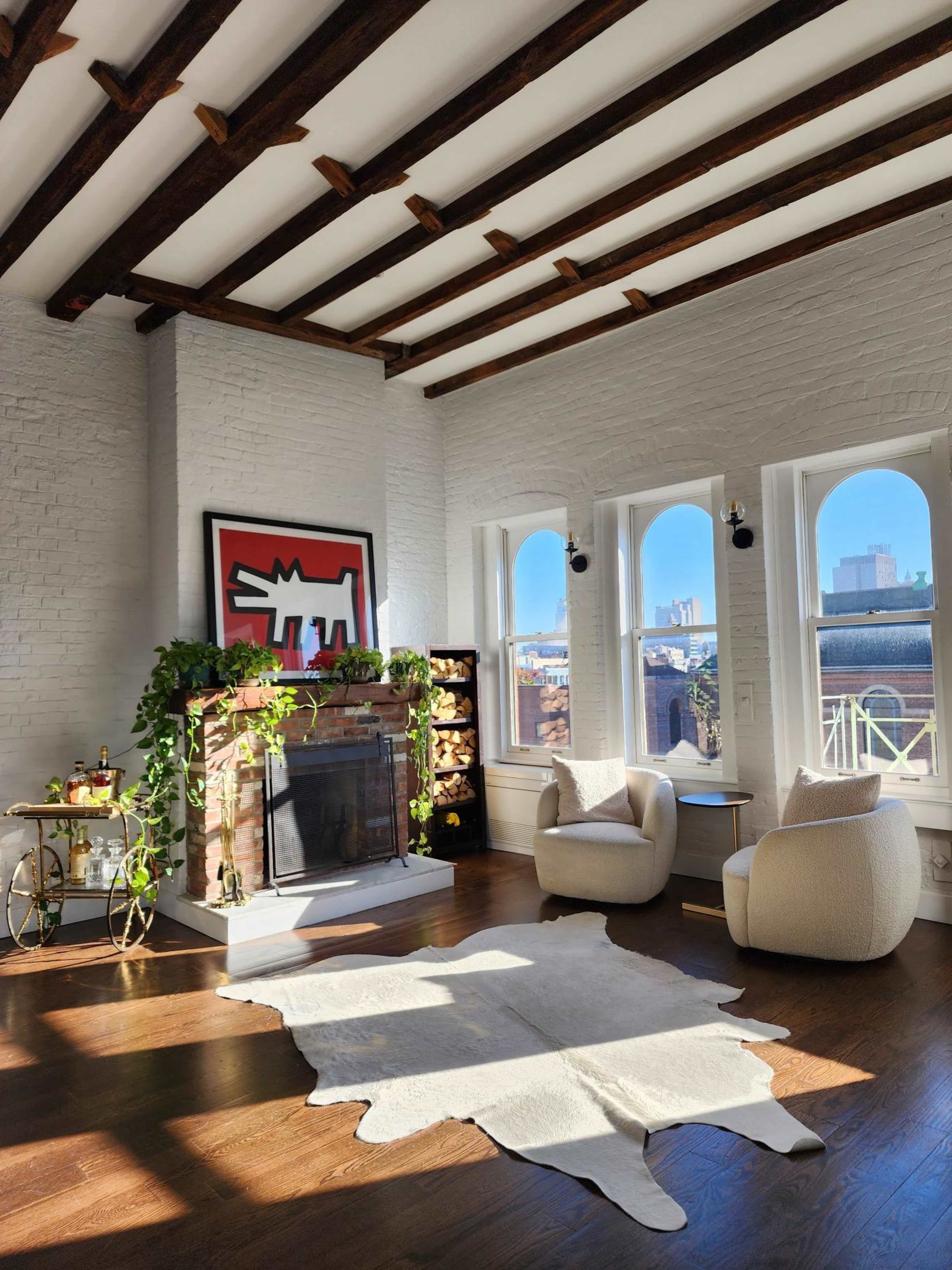 A bright living room with a fireplace, two armchairs, a bookshelf, and large windows that allow sunlight to flood the space.