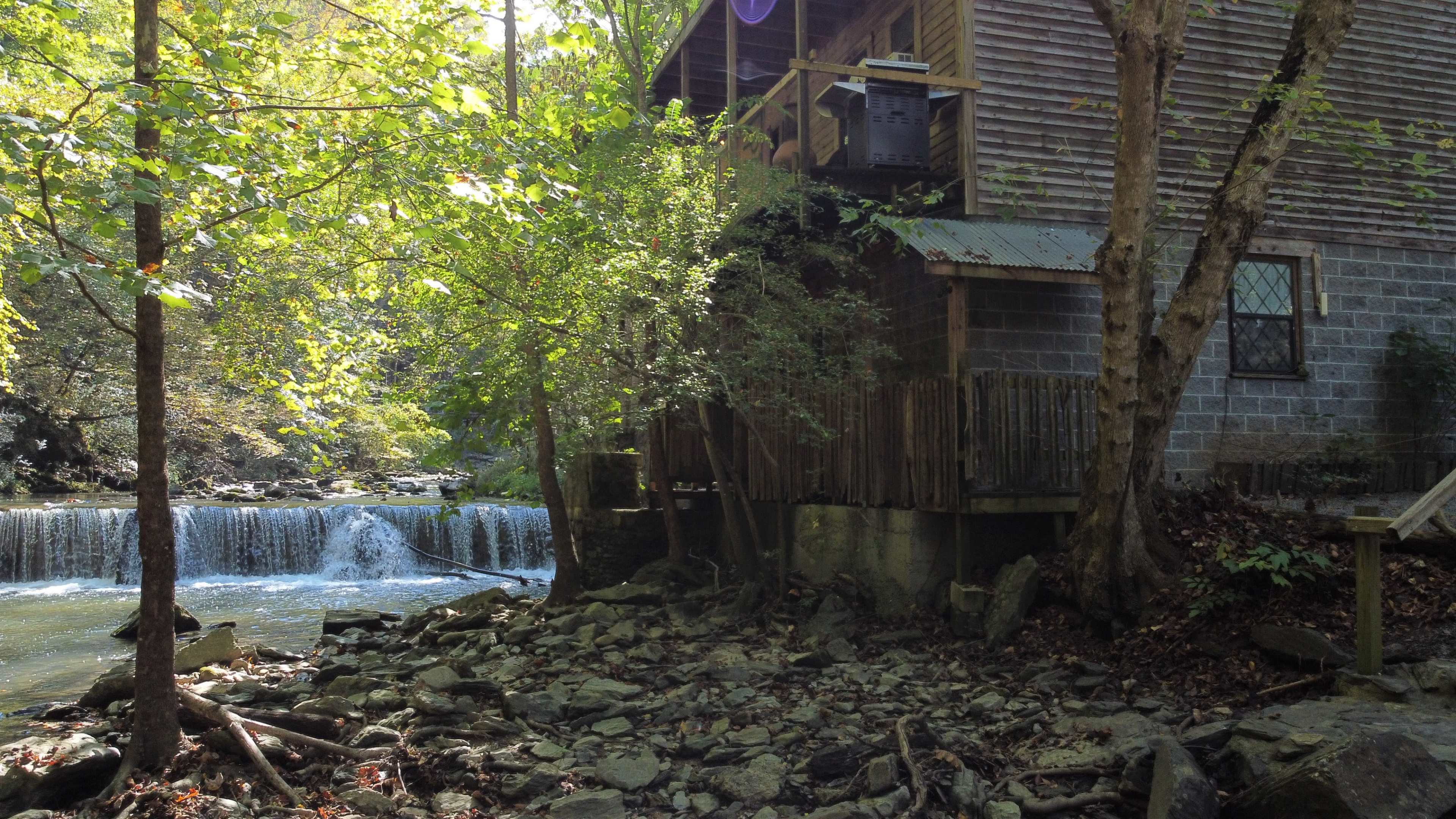 A wooden structure sits alongside a flowing river, with a small waterfall visible in the background amidst lush greenery.