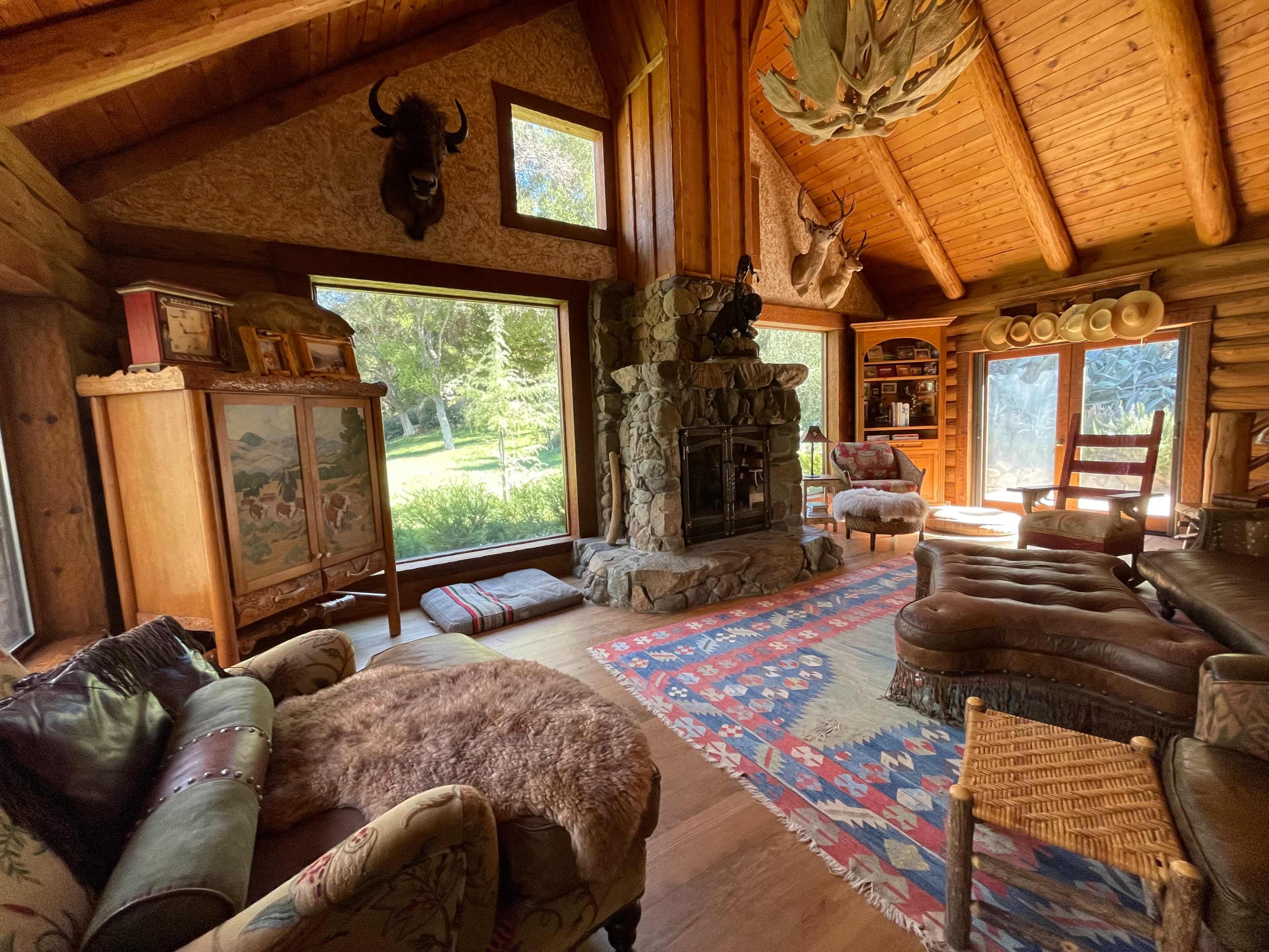 The interior of a cozy cabin features a stone fireplace, large windows overlooking a green landscape, and rustic furniture arranged around a colorful rug.