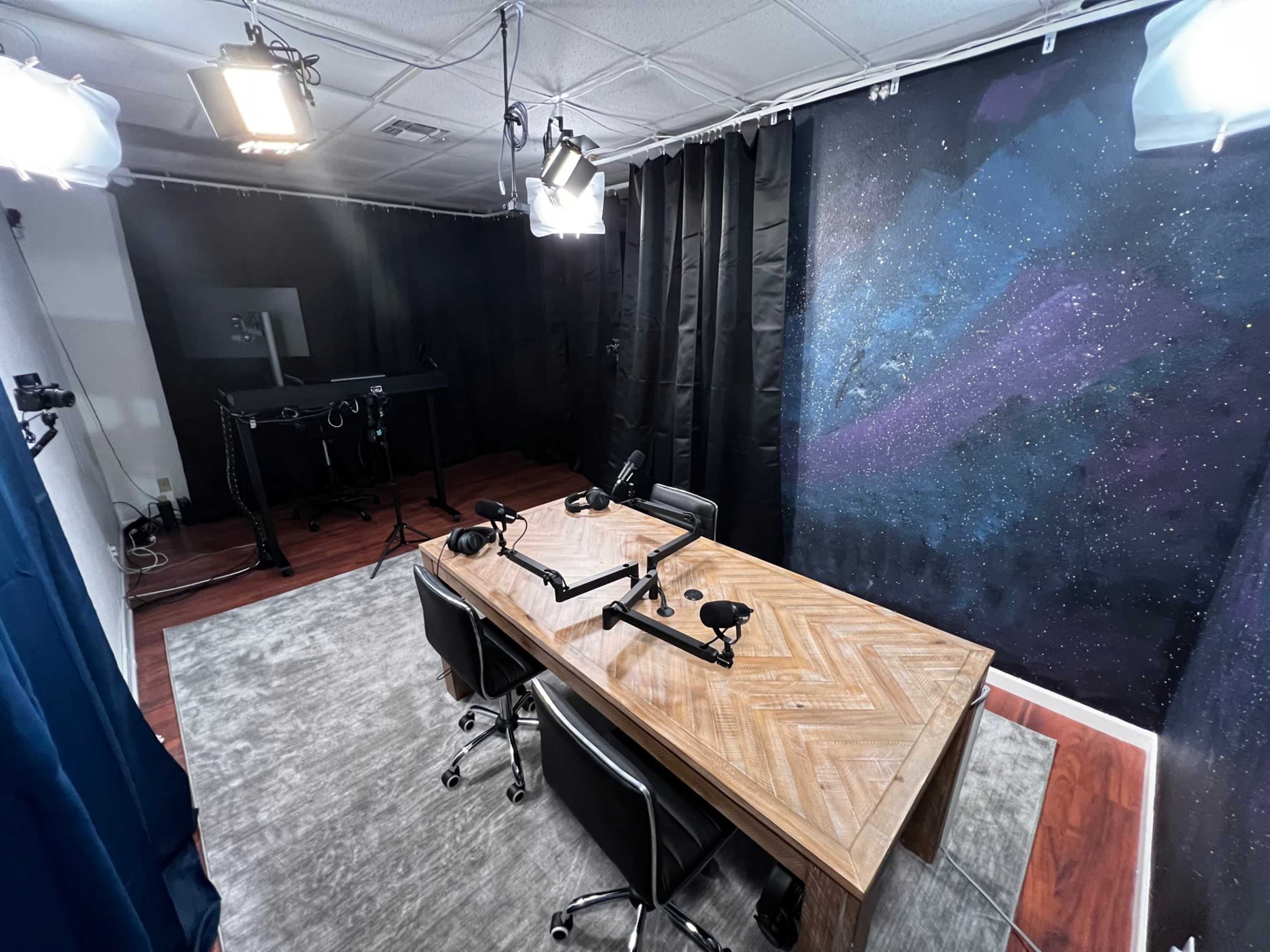 The image shows a podcast studio with a wooden table, two microphones on adjustable arms, and a starry backdrop, all under bright studio lights.