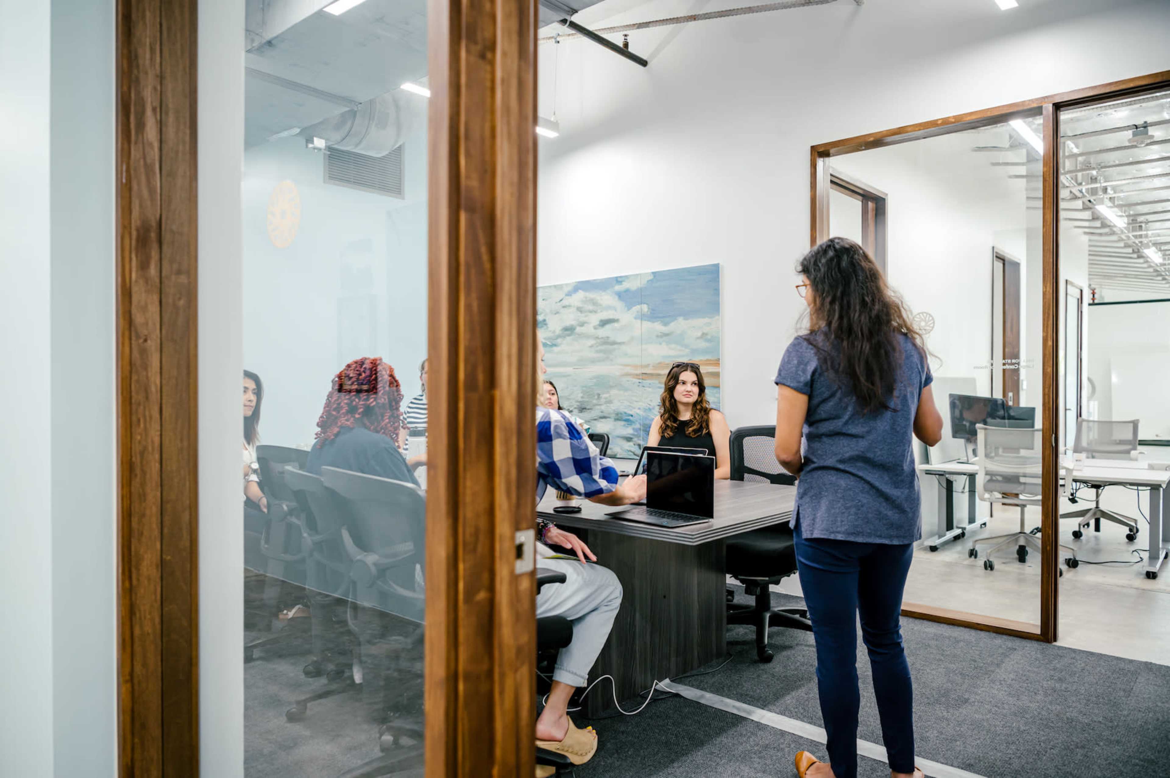 A woman stands in front of a glass-walled conference room where several people are seated at a table.