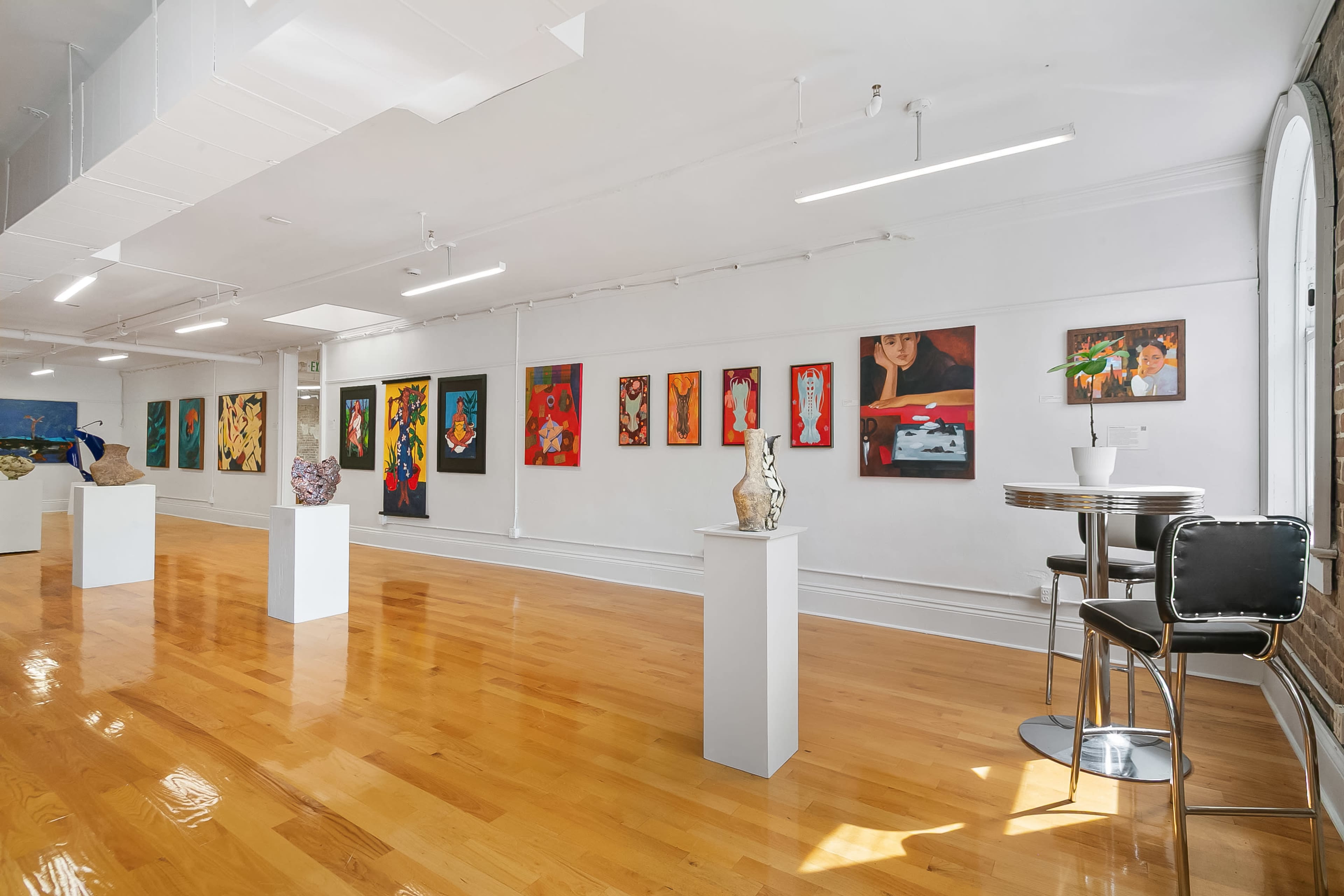 The image shows an art gallery with colorful paintings displayed on the walls and sculptures placed on white pedestals, accompanied by a small round table and chairs in the foreground.