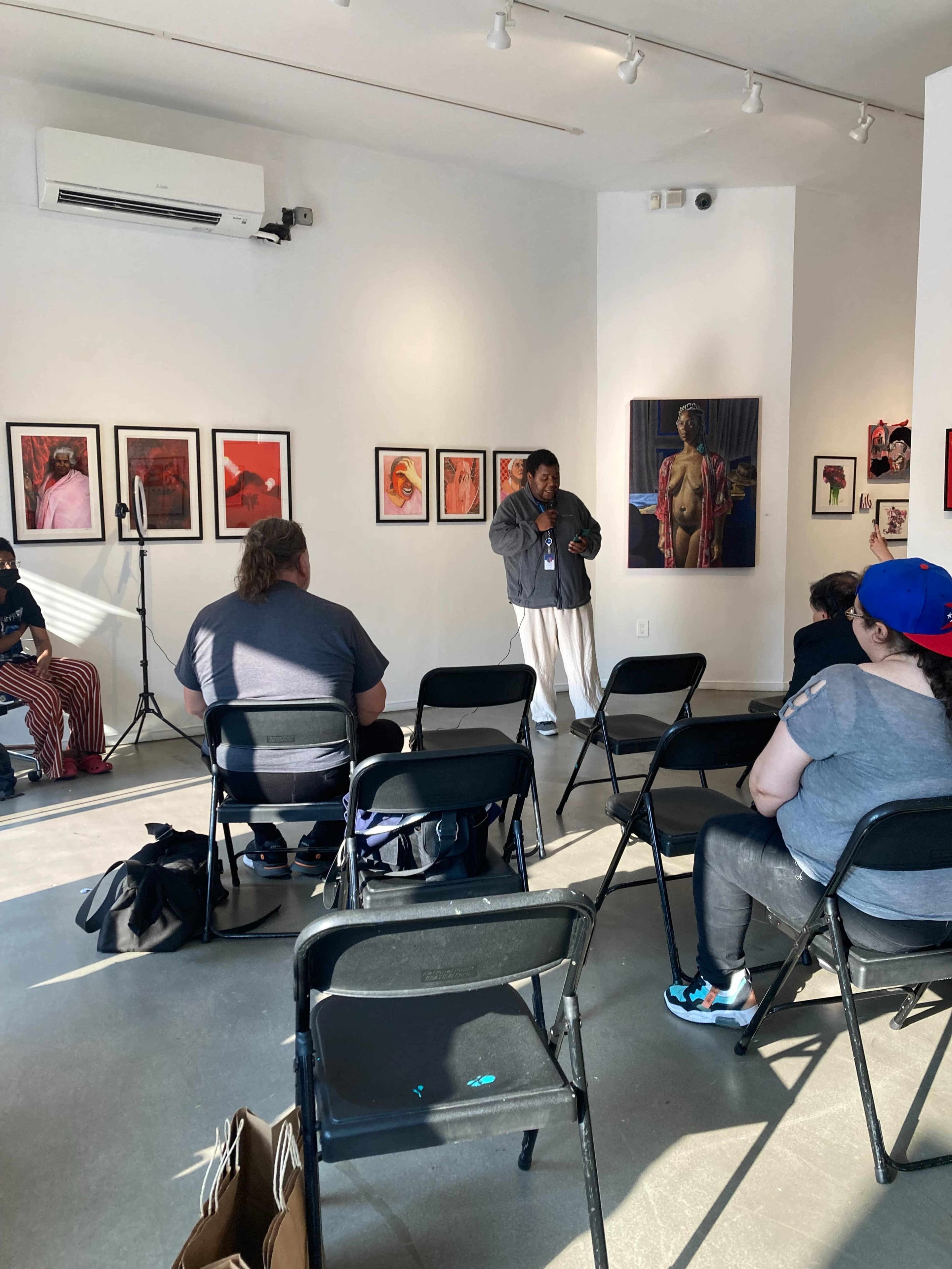 A speaker stands in front of a small audience seated on folding chairs in an art gallery surrounded by framed artworks.