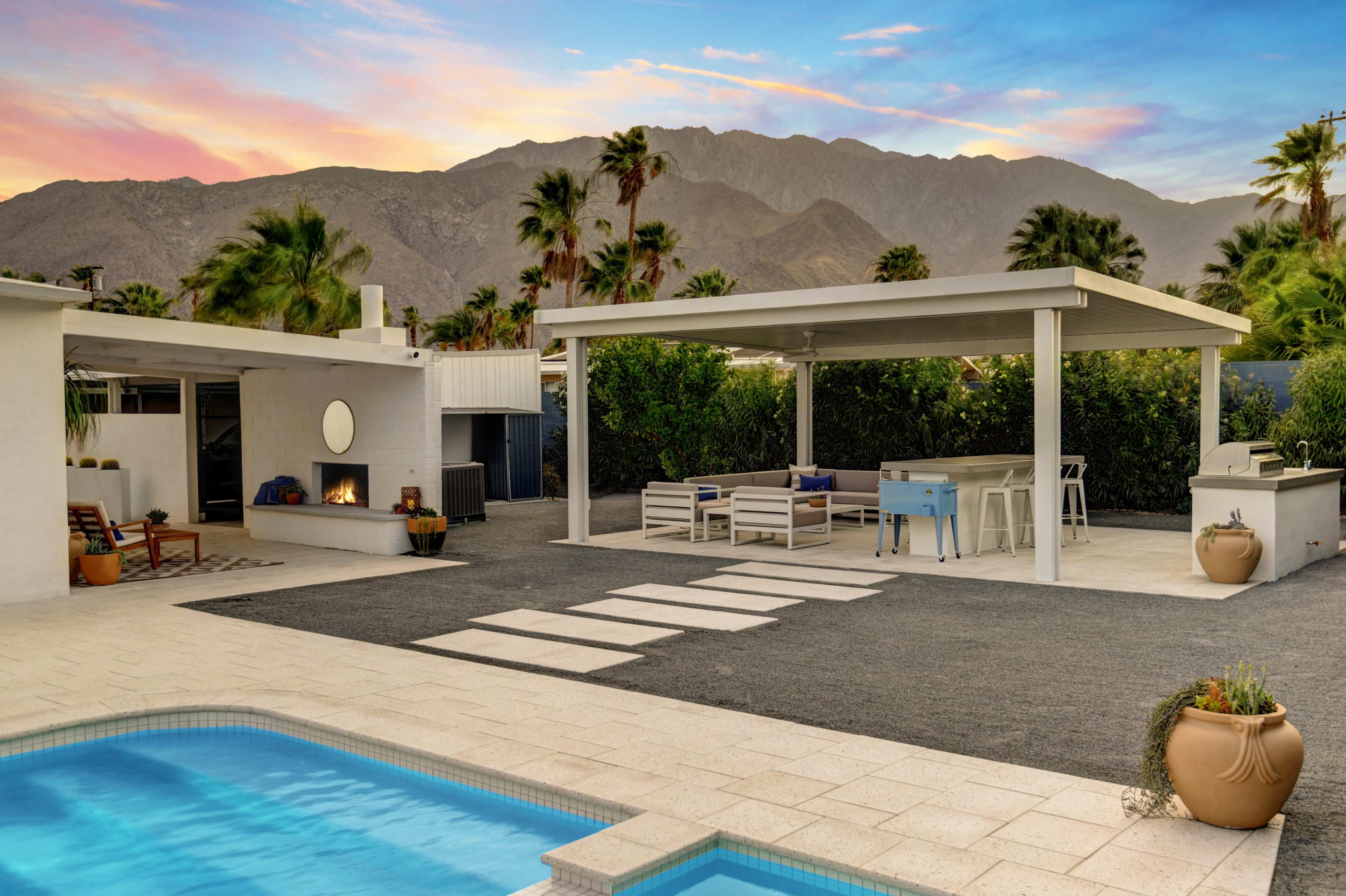 A modern outdoor space featuring a pool, a covered seating area, and a backdrop of mountains under a colorful sky.