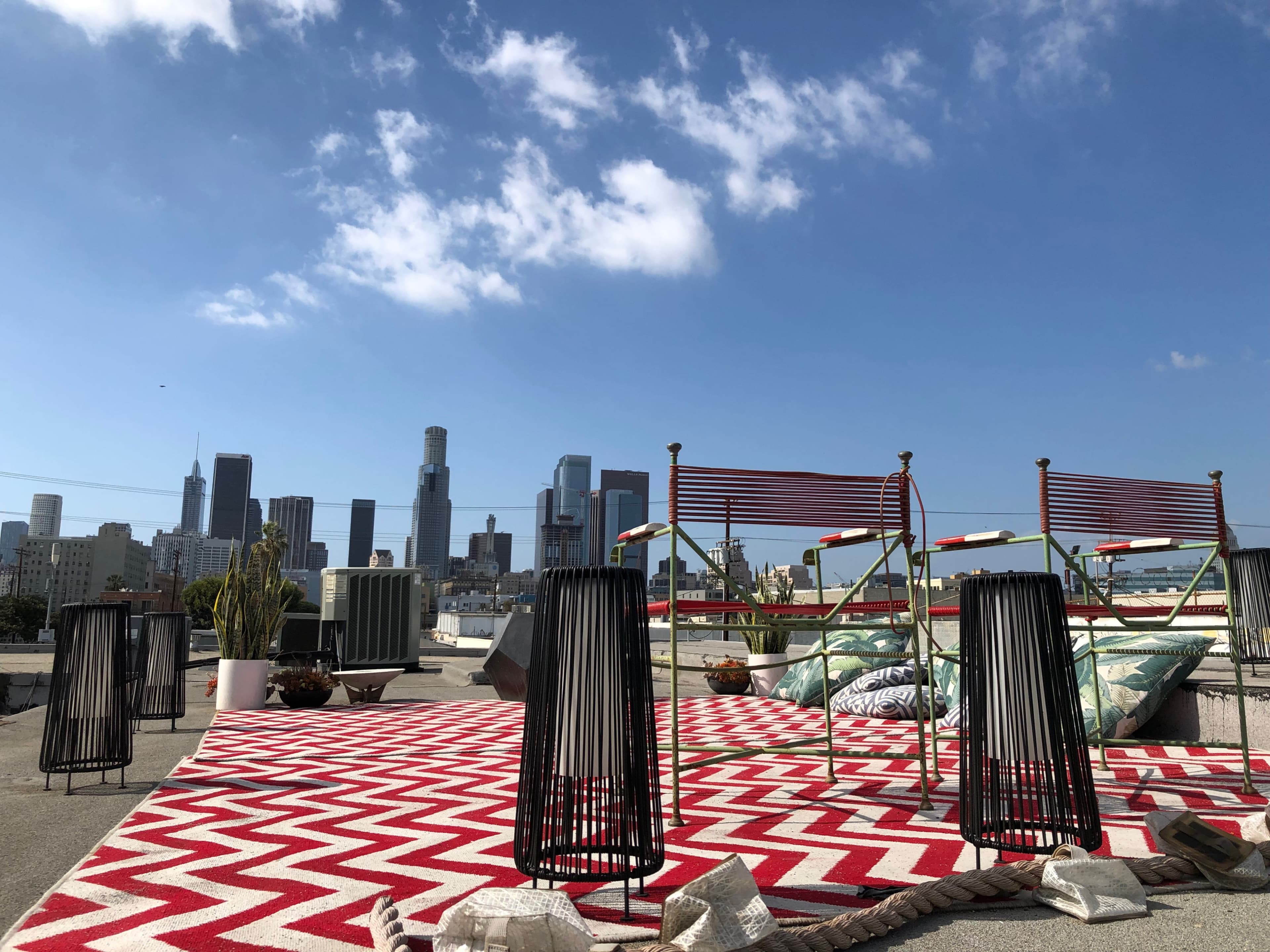 A rooftop space features a red and white patterned rug with chairs and potted plants, overlooking the skyline of Los Angeles under a blue sky.