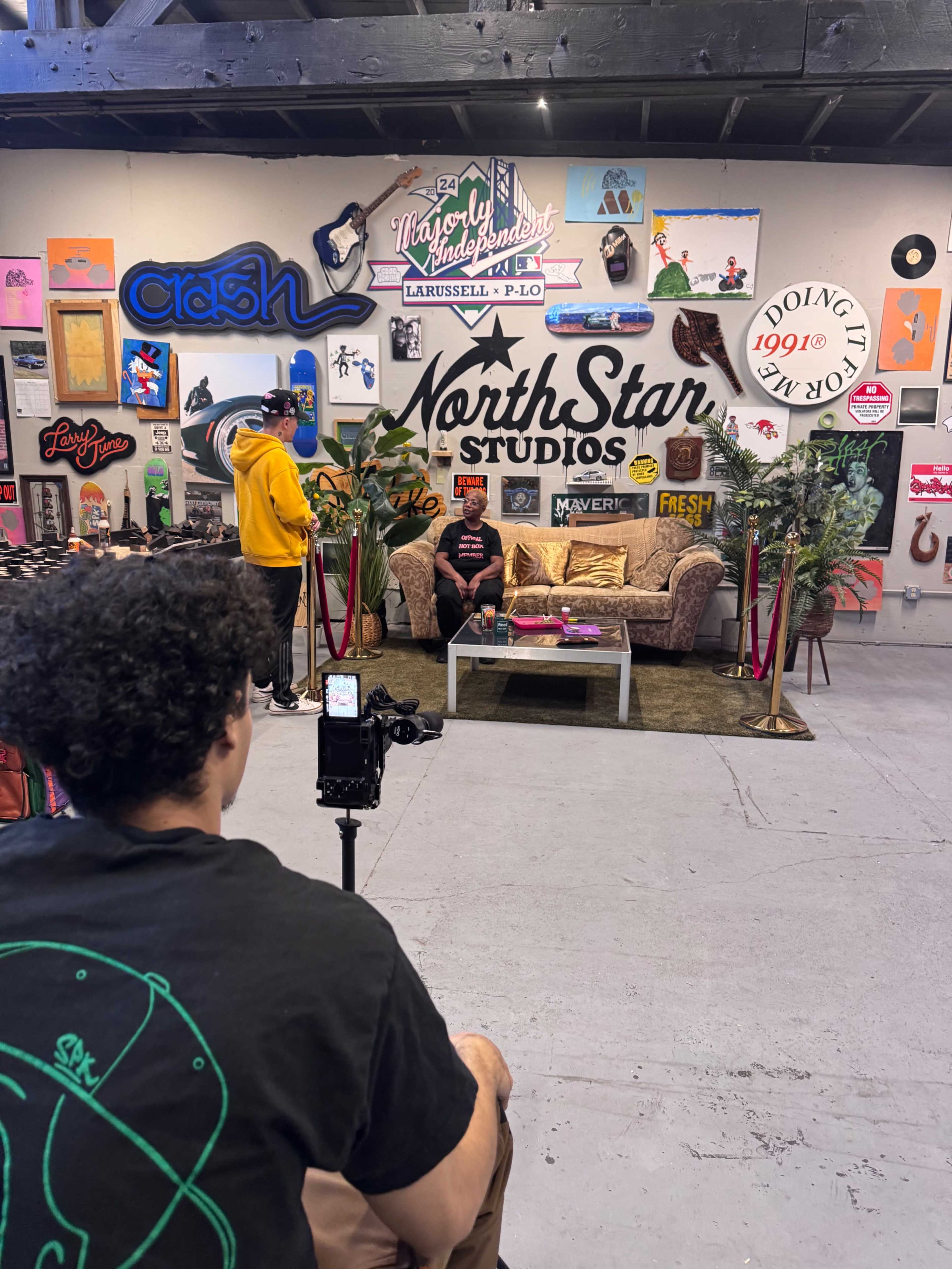 A person is seated on a couch in a studio filled with various colorful wall art, while another person is positioned behind a camera.