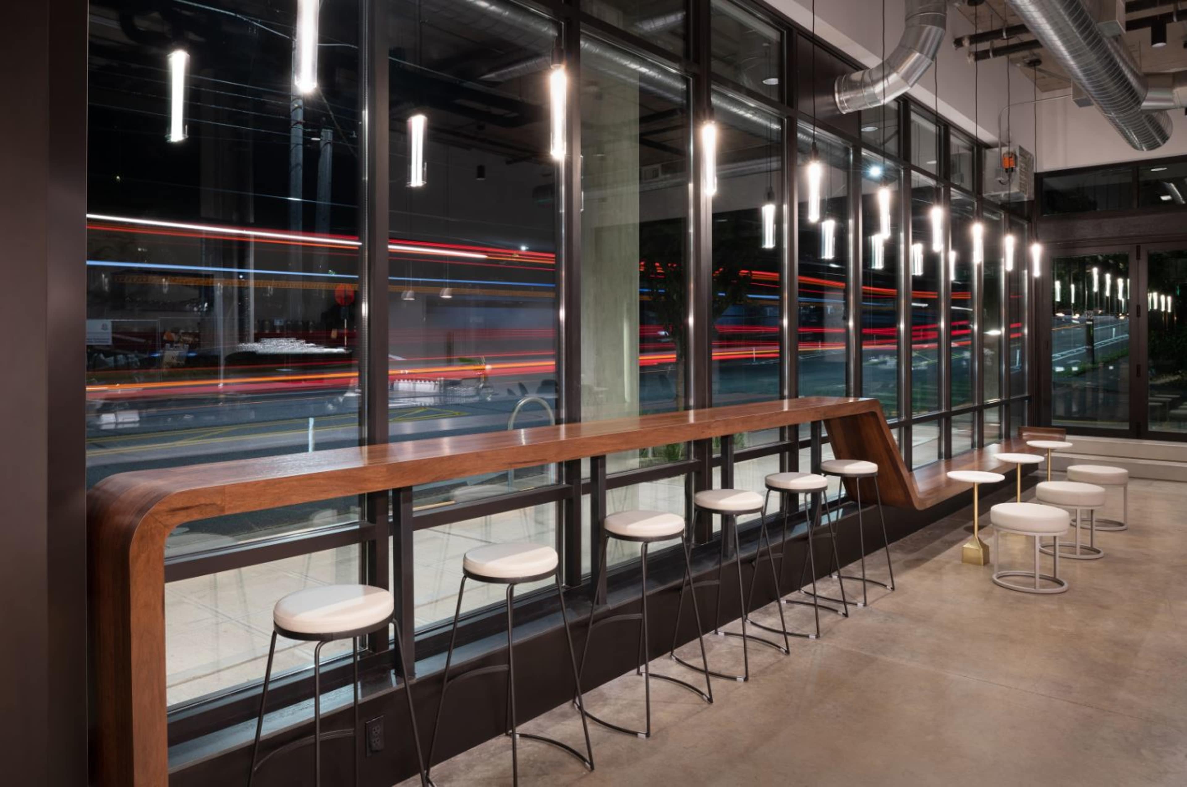 A modern café interior with a wooden counter and white bar stools, alongside large windows displaying blurred lights from passing traffic.